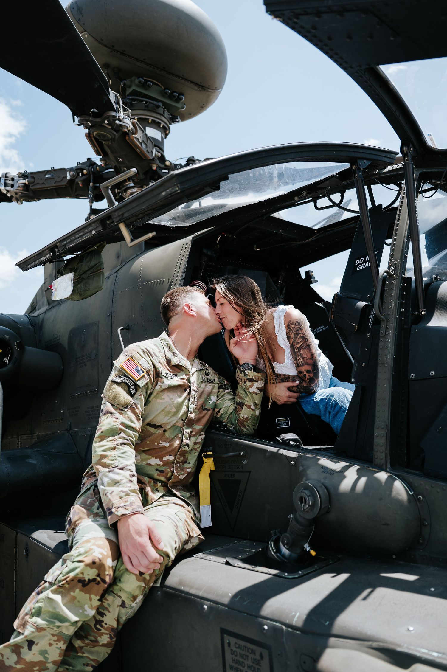 Soldier in camouflage uniform kissing a woman sitting in an Apache military helicopter cockpit outdoors.
