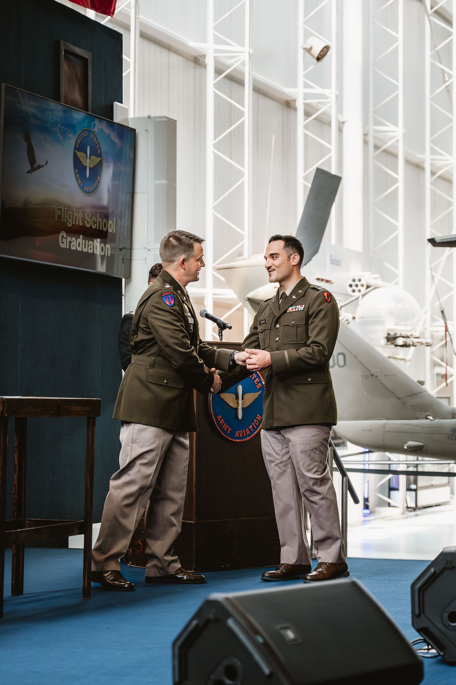 Two military officers in olive uniforms shake hands at a Flight School Graduation ceremony inside an aviation museum.