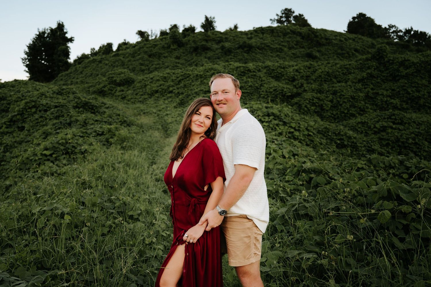Couple posing on a lush green hillside, woman in red dress and man in white shirt holding hands outdoors.