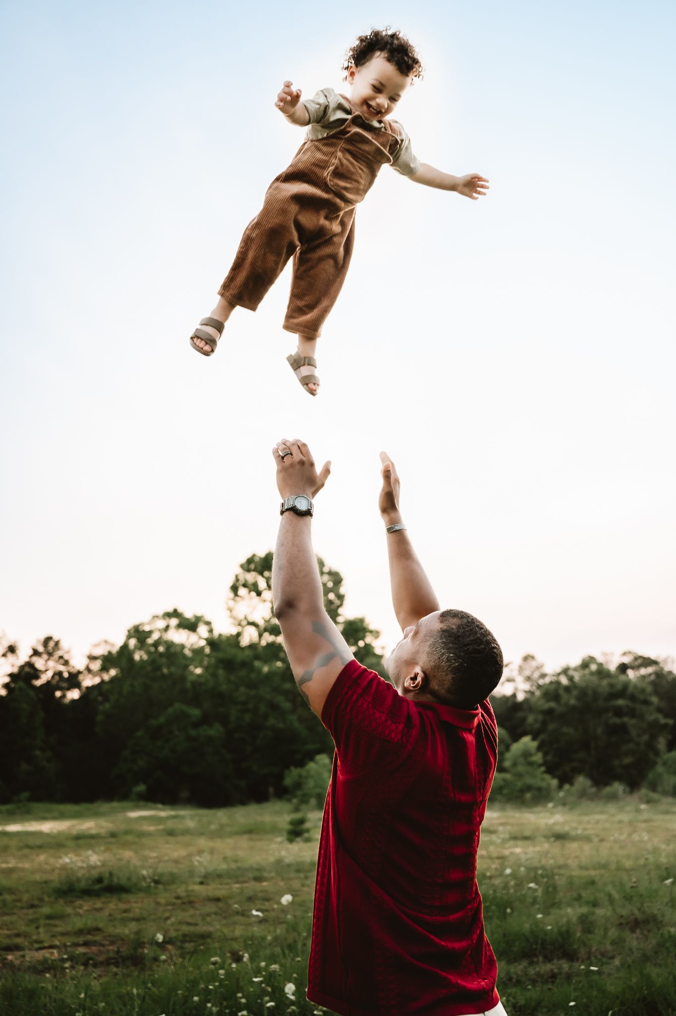 In a sunlit field, someone in a burgundy shirt throws a small child wearing brown overalls joyfully into the air.