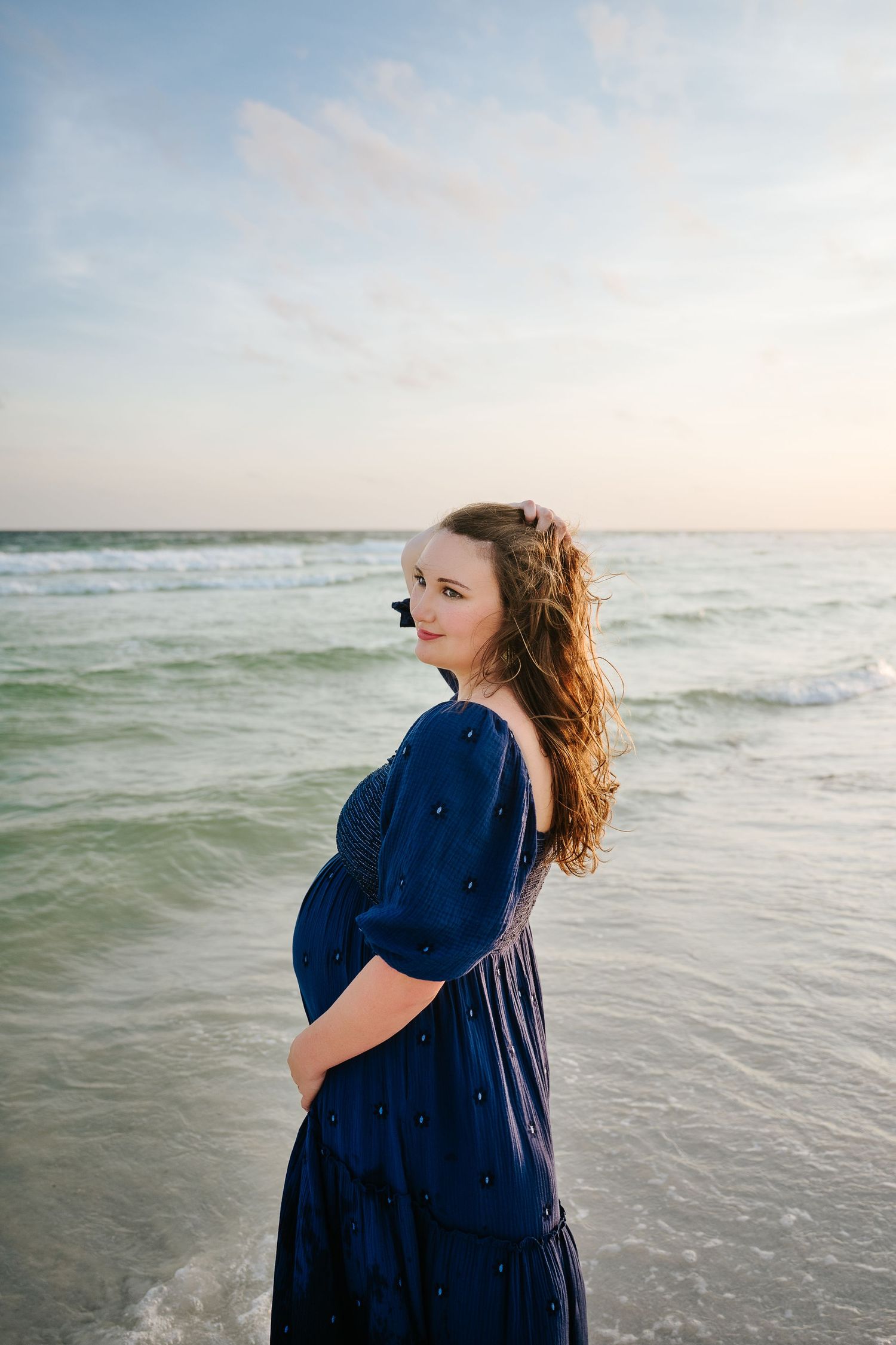 Pregnant woman in navy dress cradling baby bump on beach at sunset with ocean waves behind her.
