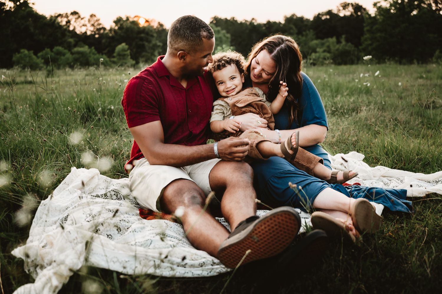 Family in casual attire relaxes together on white blanket in grassy field during golden sunset hour.