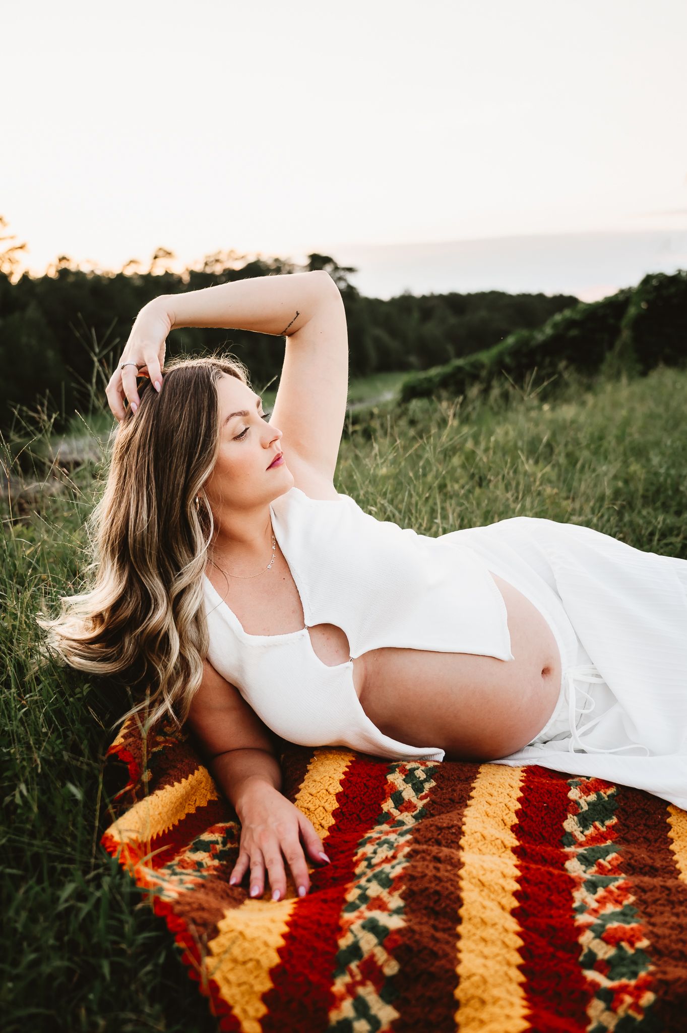 Person in white dress reclining on vintage orange and red striped blanket in sunlit meadow at golden hour.