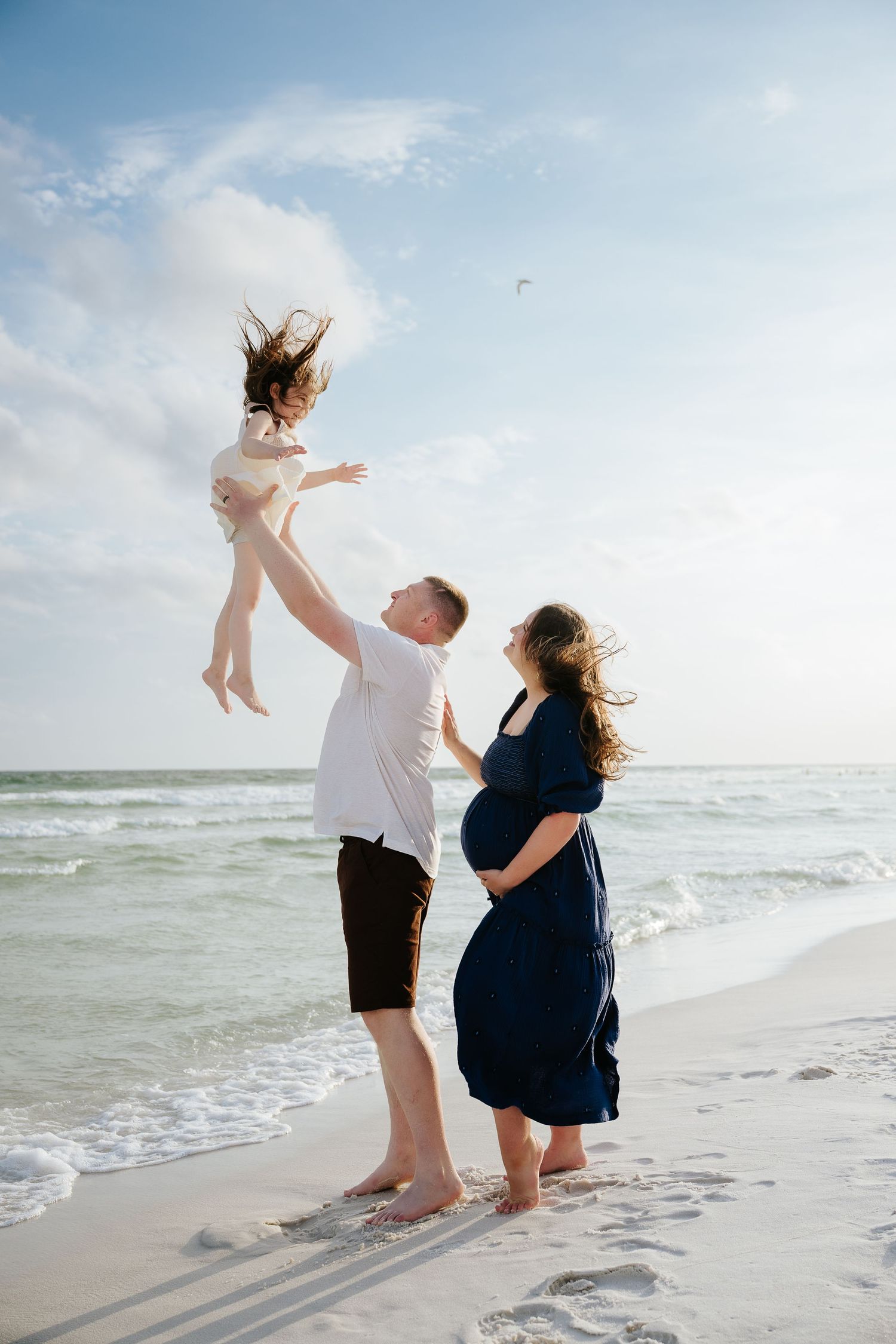 Pregnant woman in navy dress watches man toss child in air on sunny beach with ocean waves.