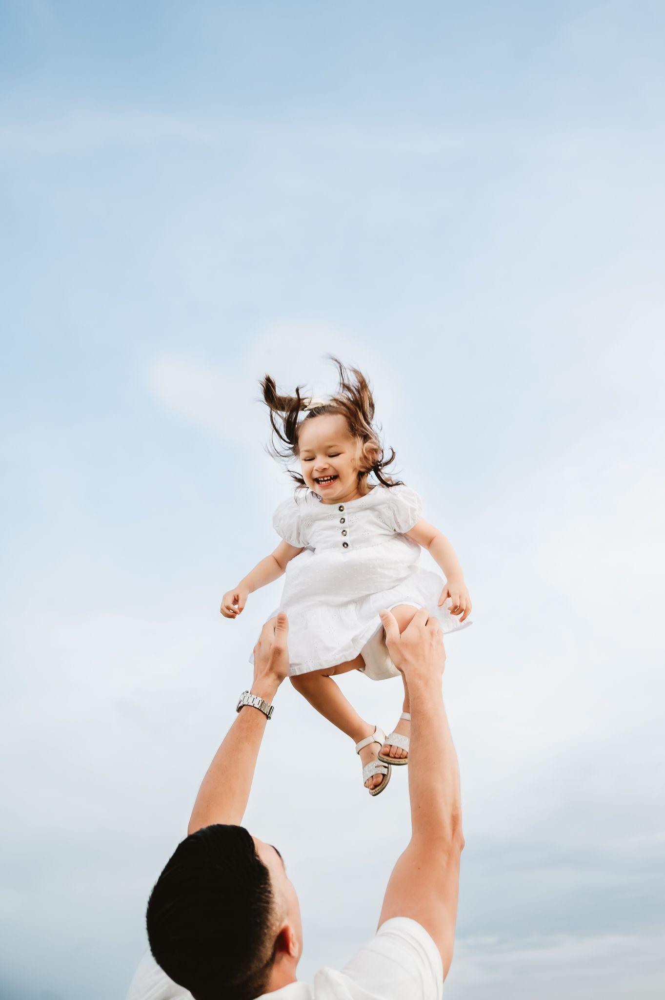 A playful outdoor scene shows a child being lifted up into the air against a bright cloudy sky.