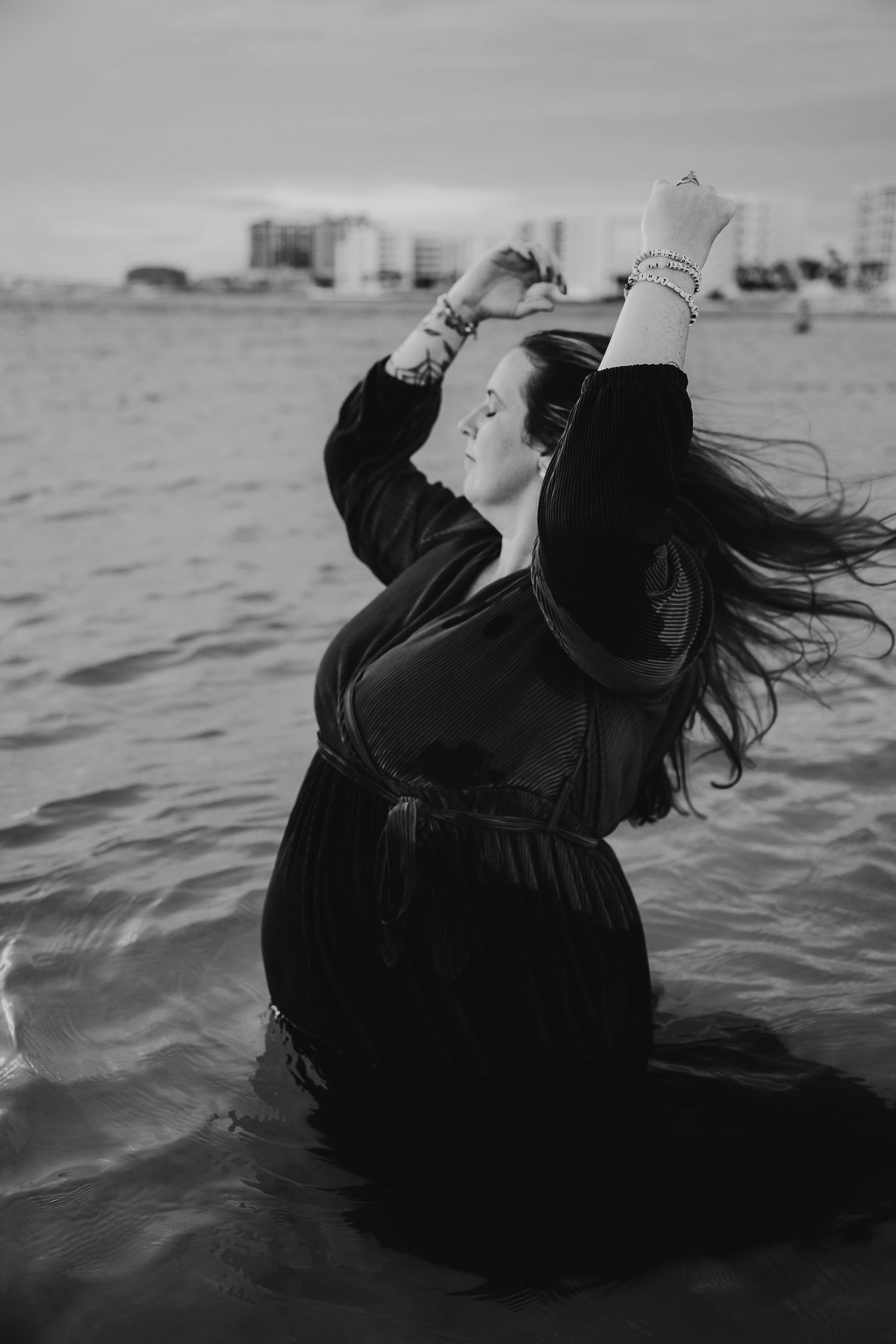 Black and white photo of a woman in a dark dress standing in ocean water with arms raised and hair blowing in the wind.