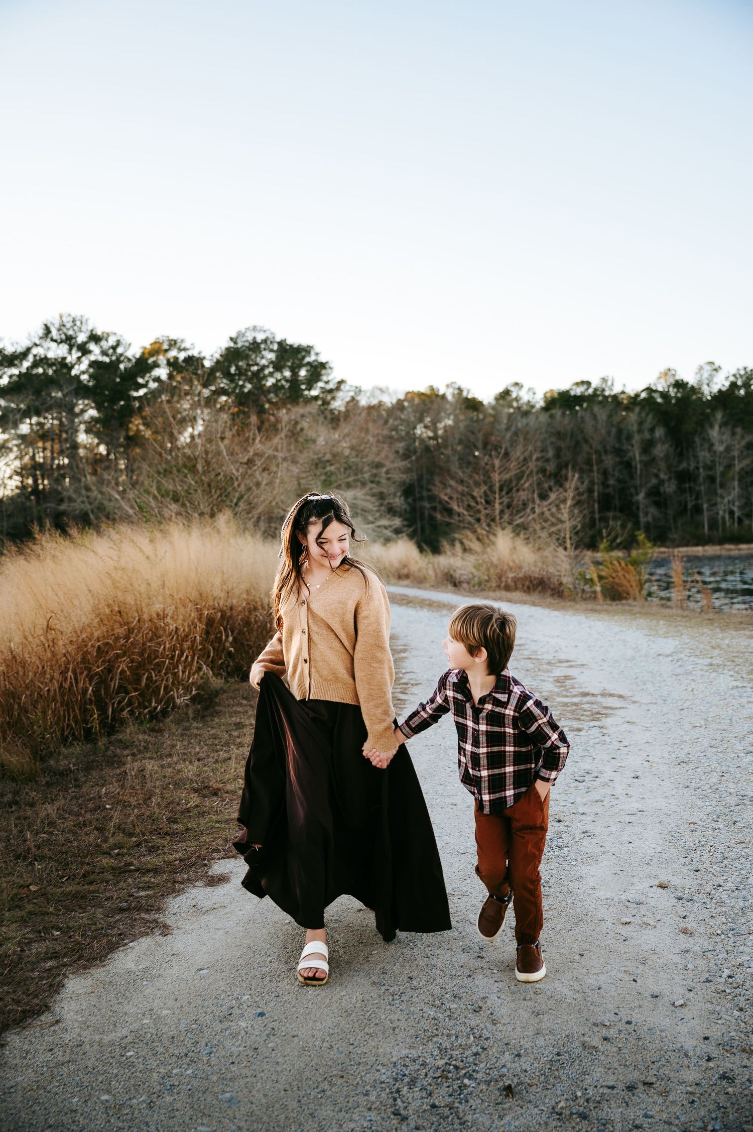 A parent and child walk hand in hand down a nature trail surrounded by tall grass and trees at sunset.
