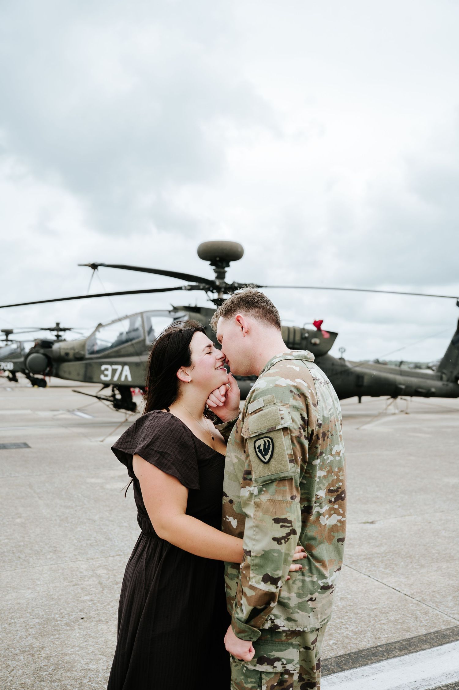 A soldier in camouflage uniform shares a romantic moment with a woman in a black dress near military helicopters.