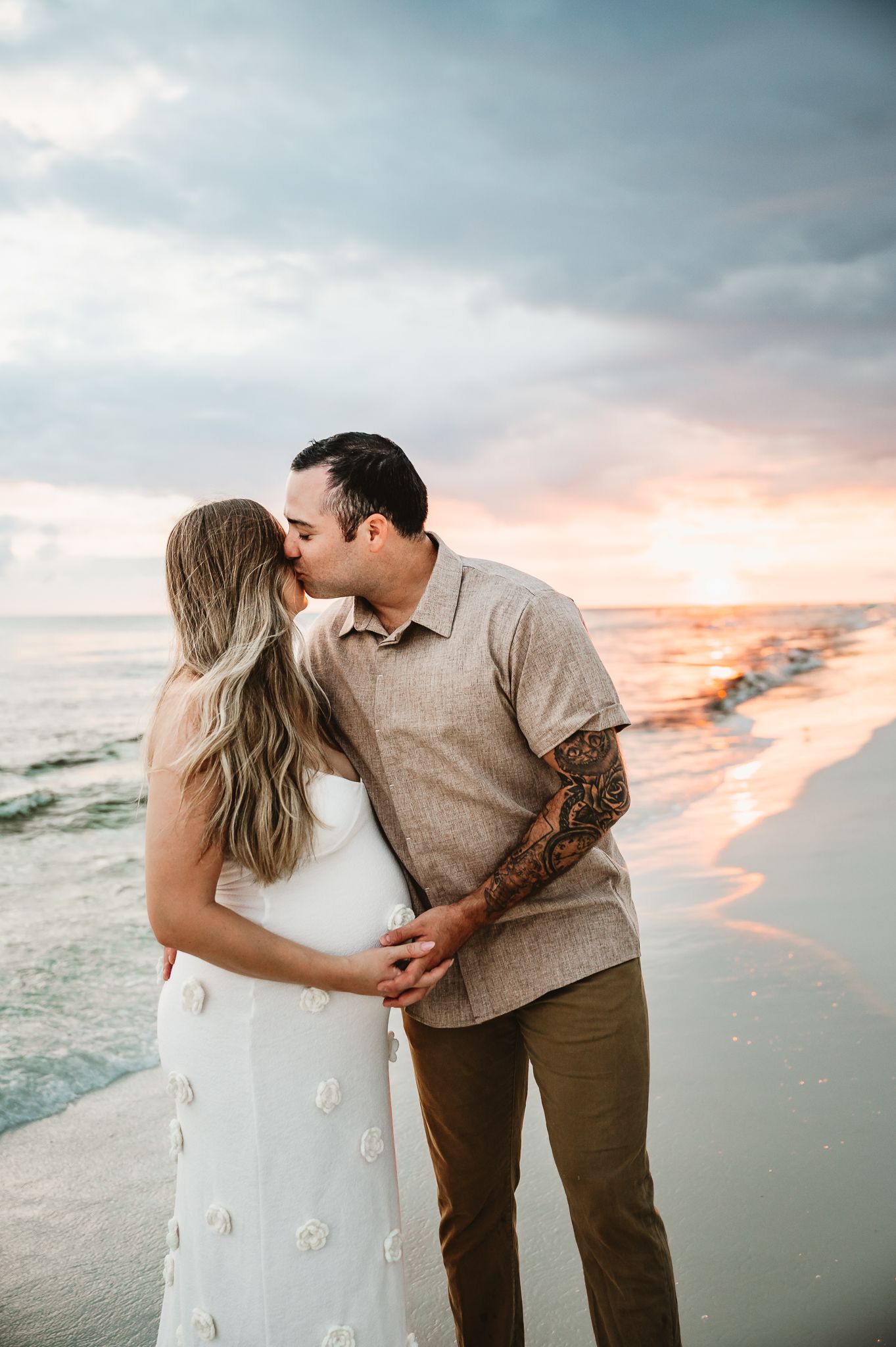 A couple shares a romantic kiss on the beach at sunset as waves lap at the shoreline.