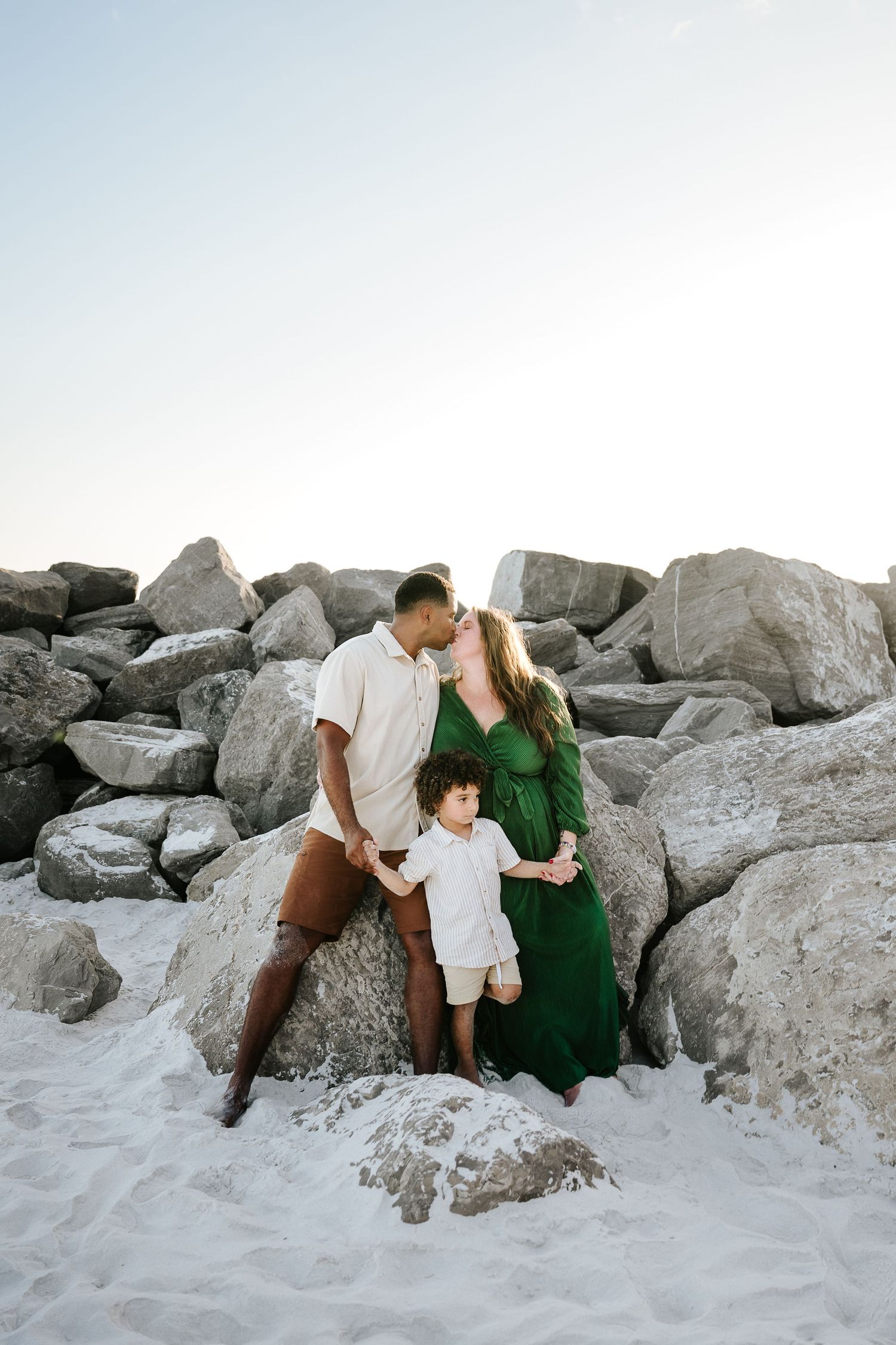 Pregnant woman in green dress kisses man while toddler stands between them on sandy beach with rocks.