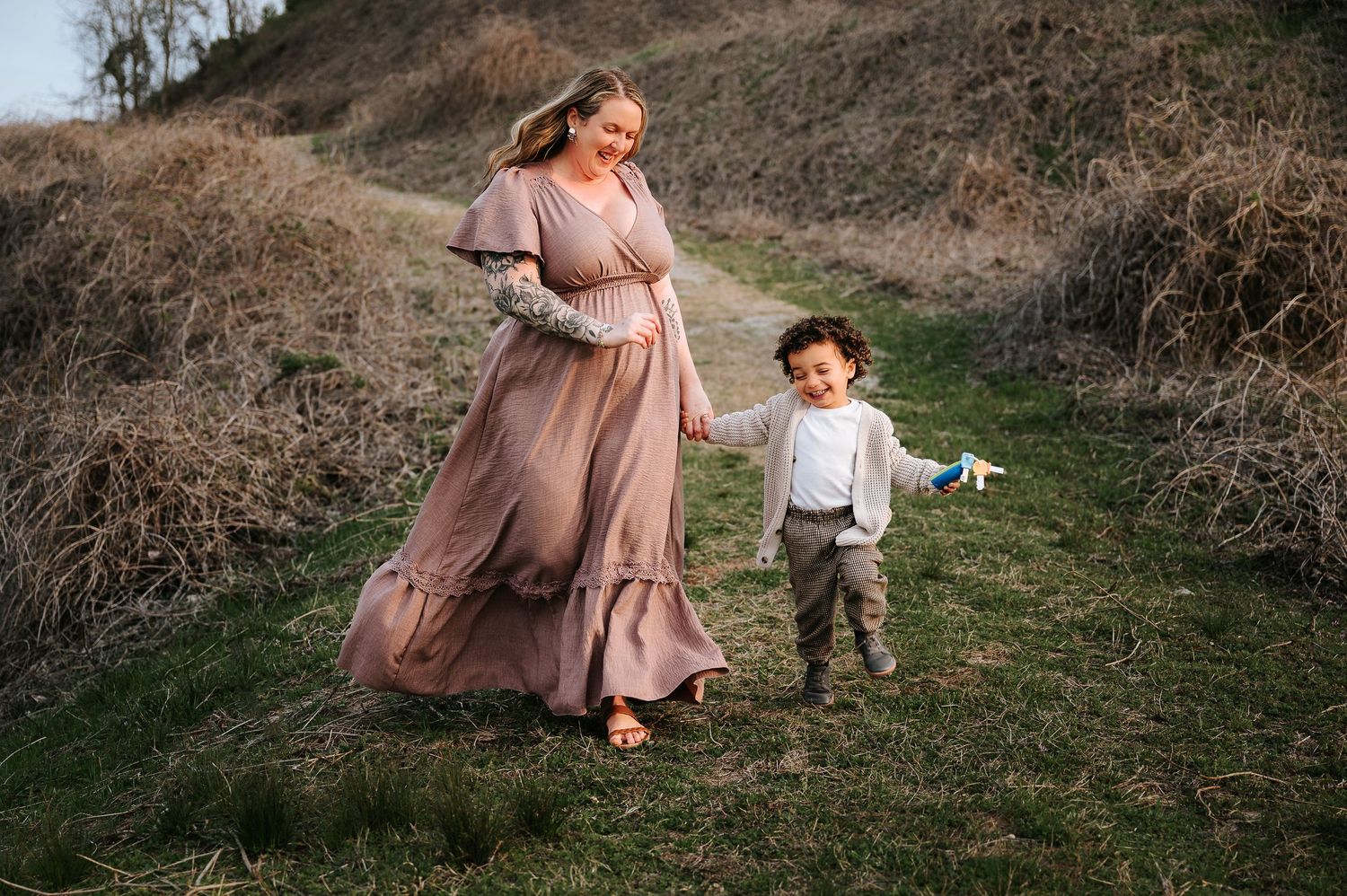 Mother and child in dusty rose dress and brown outfit play together on a grassy hillside at sunset.