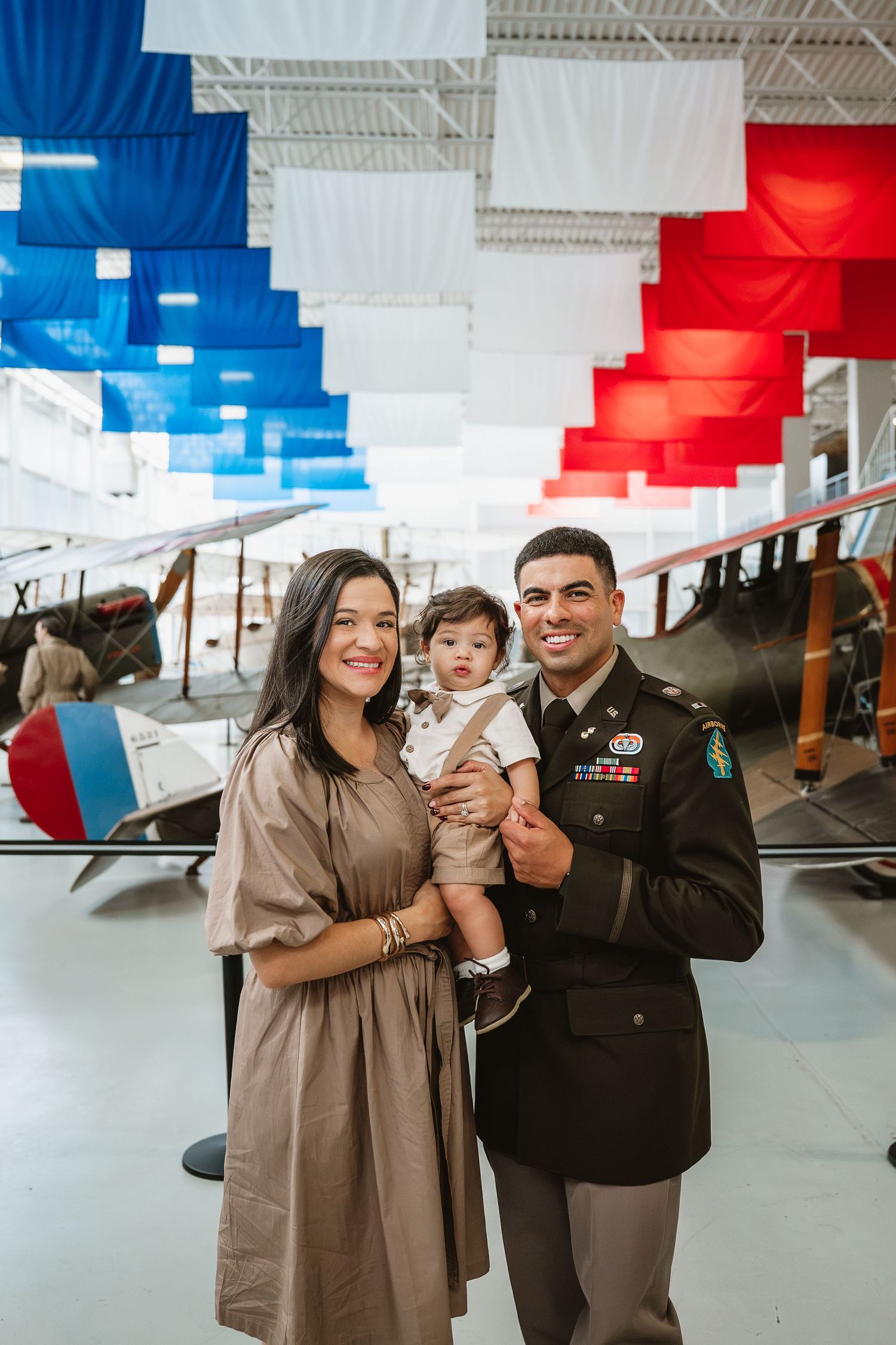Military family portrait with soldier in dress uniform holding baby beside woman in museum with patriotic banners.