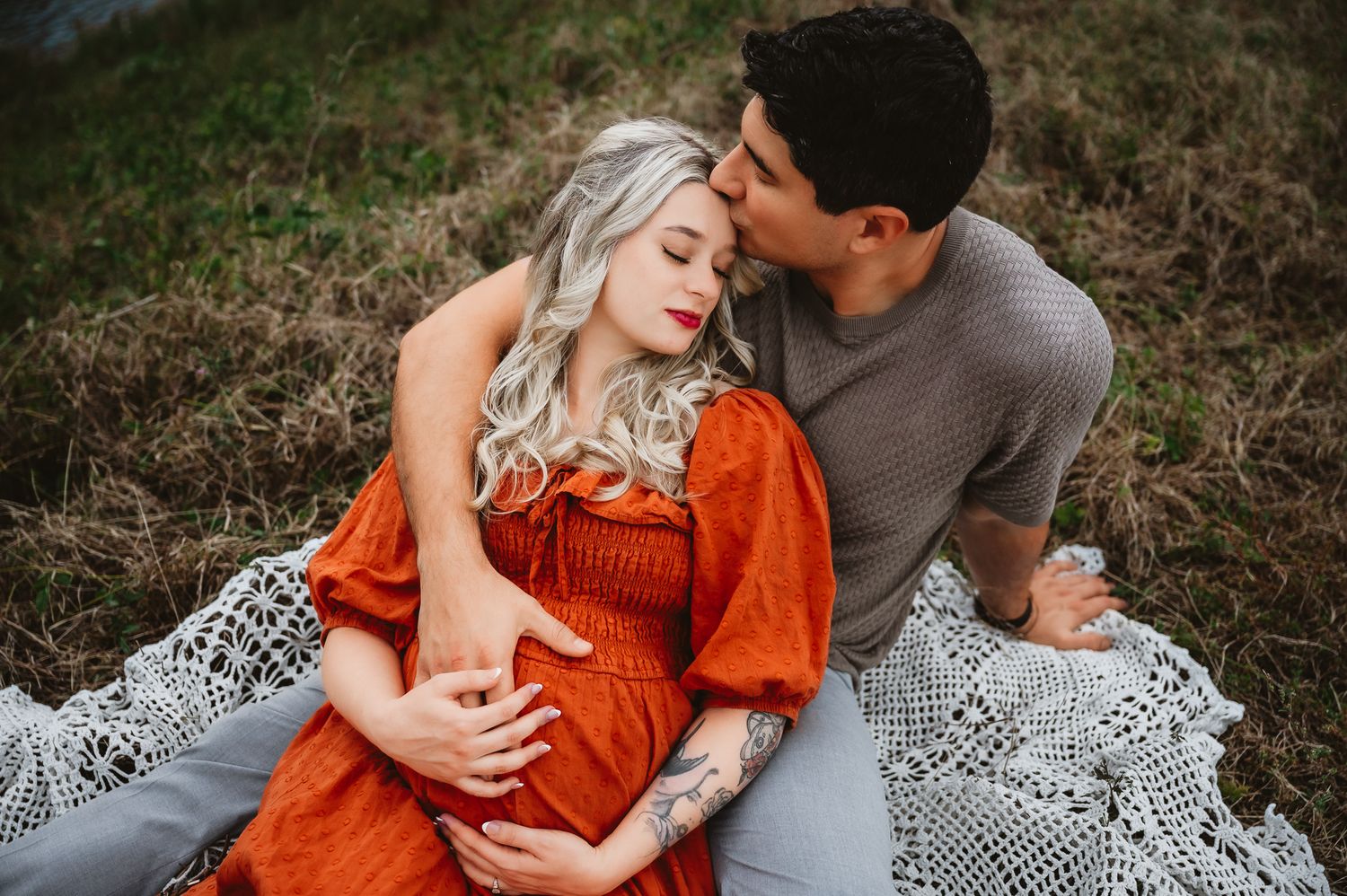 A couple shares a tender maternity moment outdoors, the woman in an orange dress cradling her baby bump.