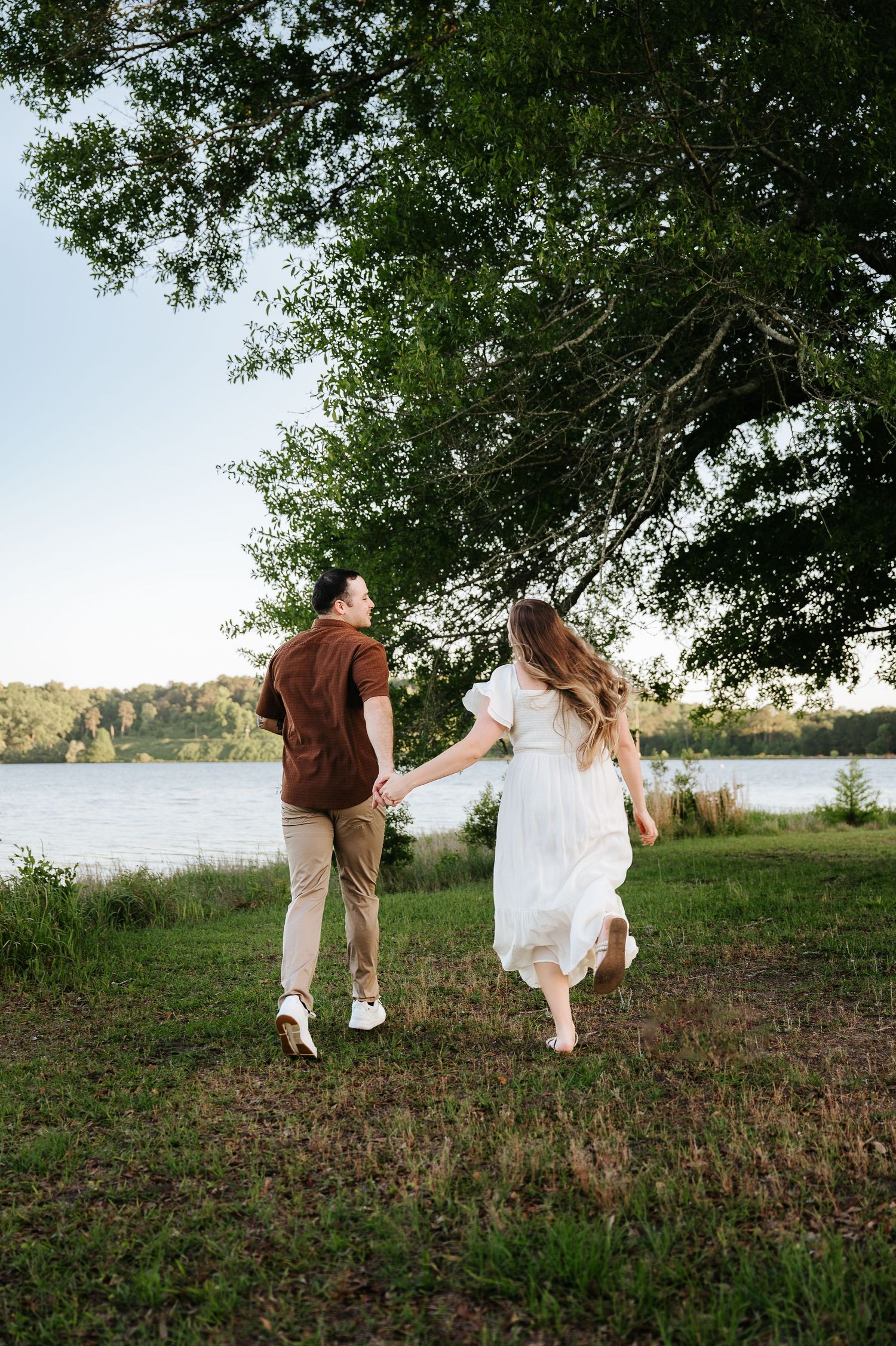 A couple holds hands and runs through a lush green park near a lake, the woman in a white dress and man in brown shirt.