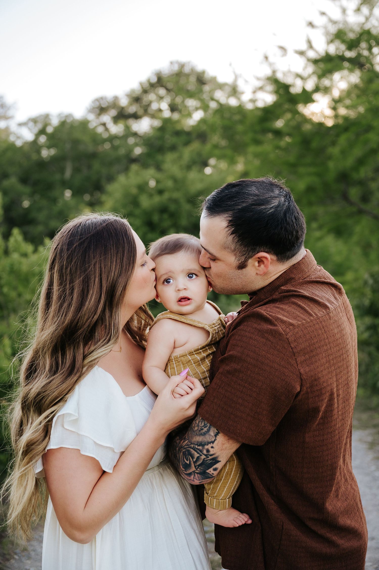 Parents kissing their baby girl on the cheeks during an outdoor family portrait session with green trees in the background.