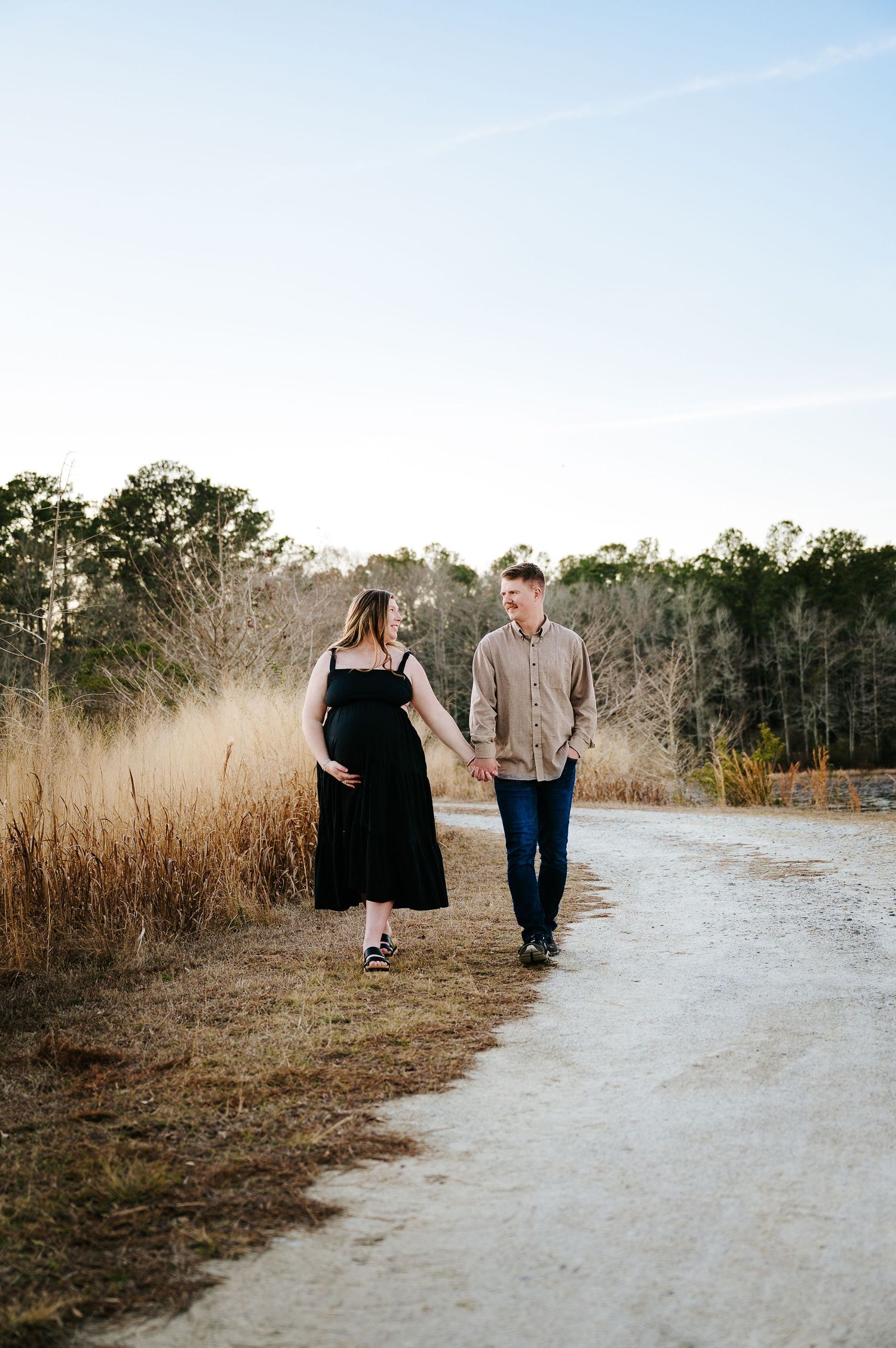 A couple walks together on a dirt path surrounded by tall dried grass and trees during golden hour.