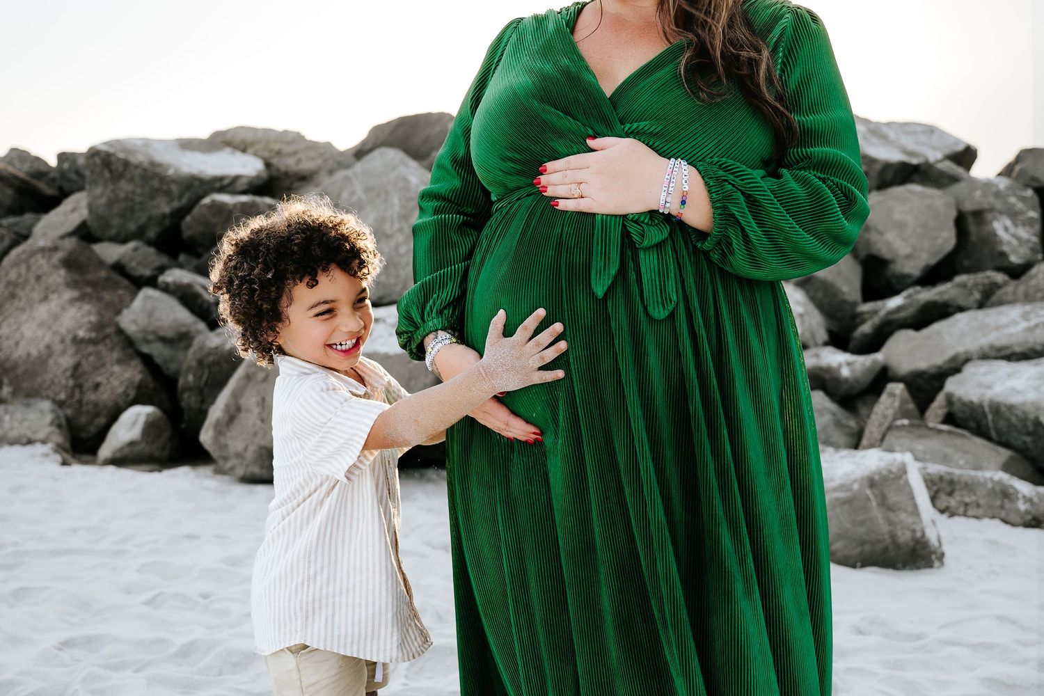 A young child touches a pregnant woman's belly outdoors near rocks, both dressed elegantly.
