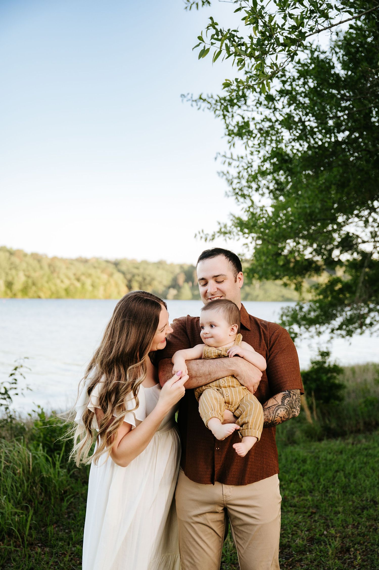 A young family poses outdoors by a lake, father holding baby while mother smiles, surrounded by lush green trees.