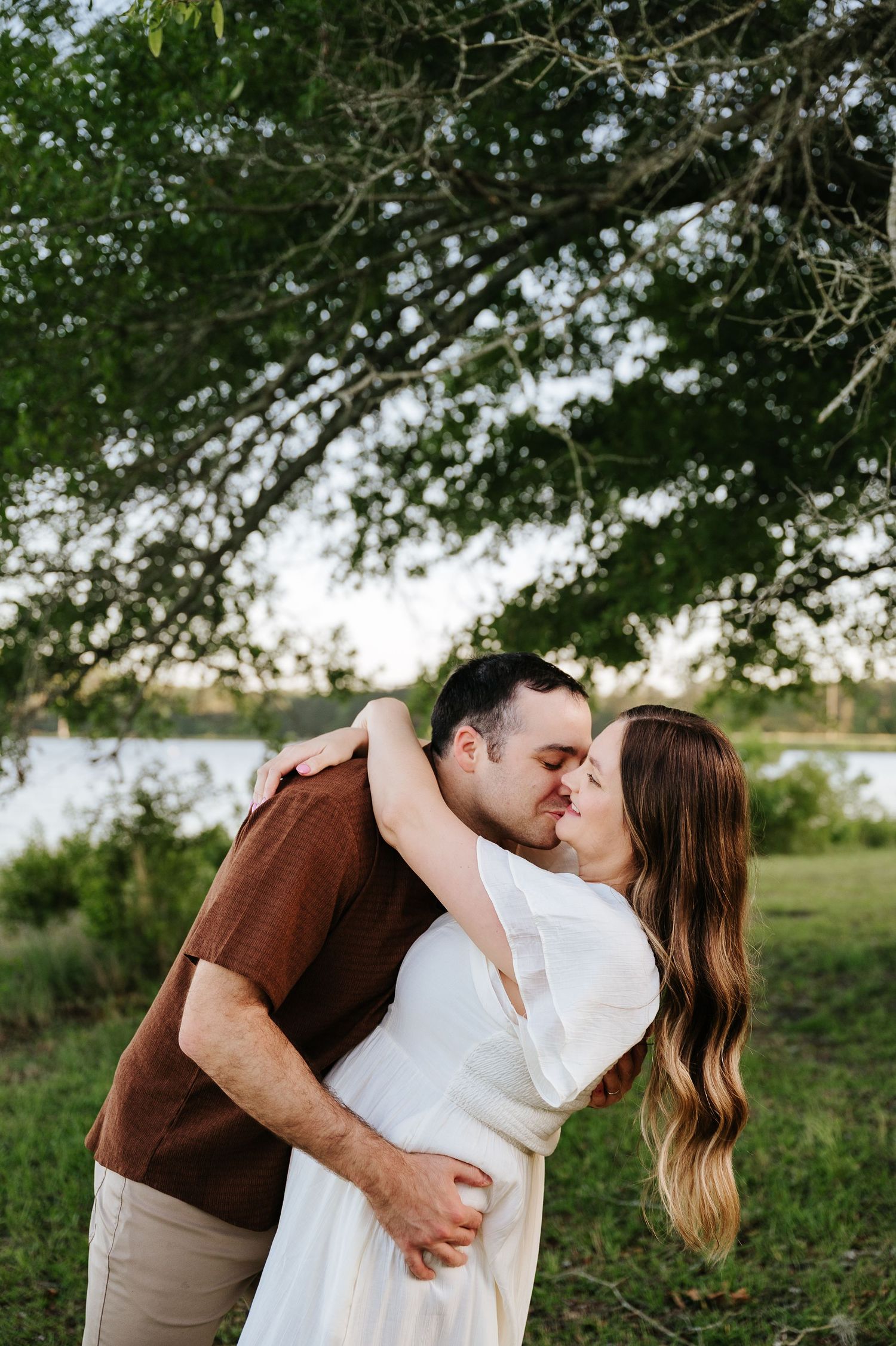 Couple sharing a romantic kiss outdoors near a lake, man in brown shirt embracing woman in white dress under a green tree.