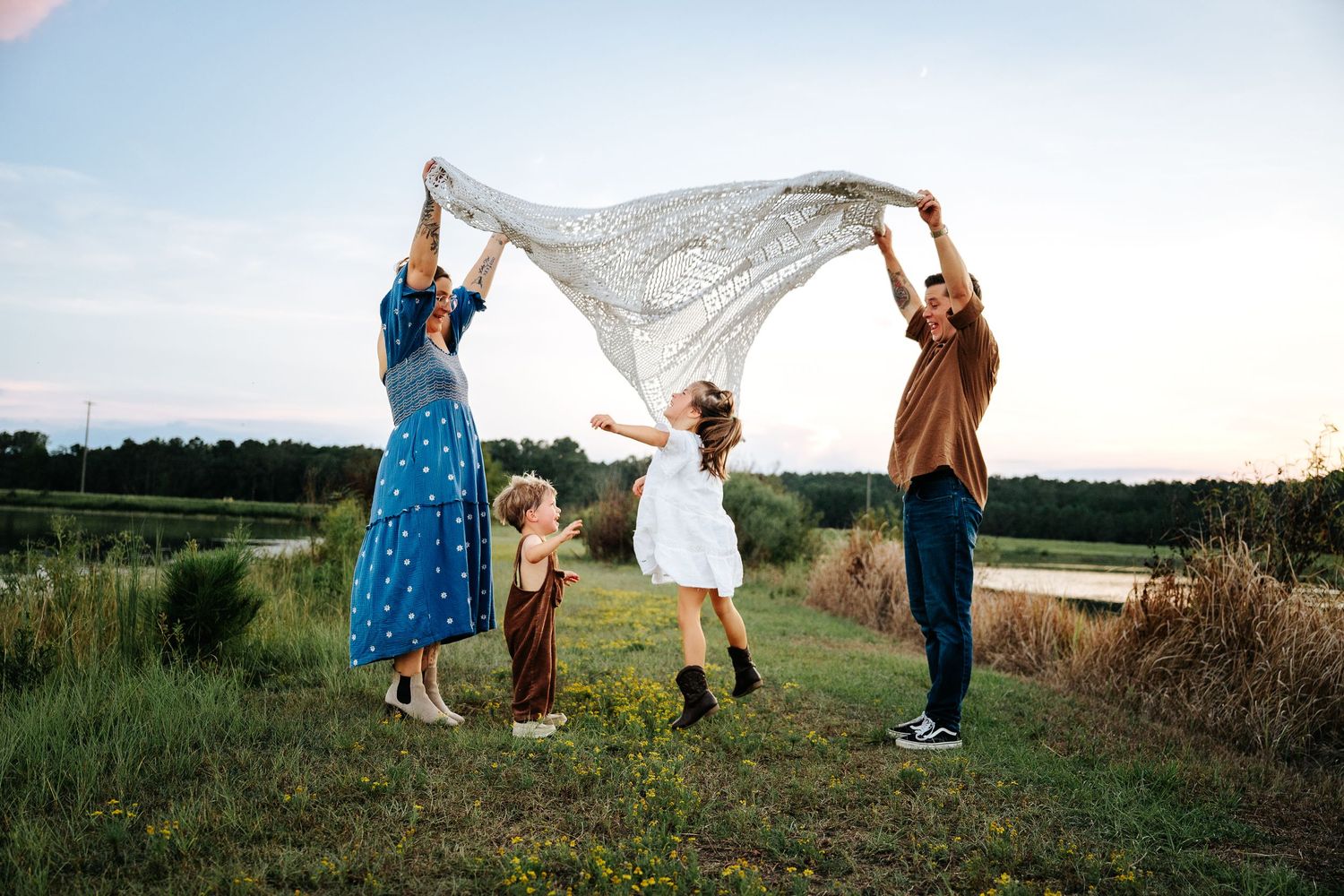 Family of four playing with a lace blanket outdoors at dusk in a green field near a pond.