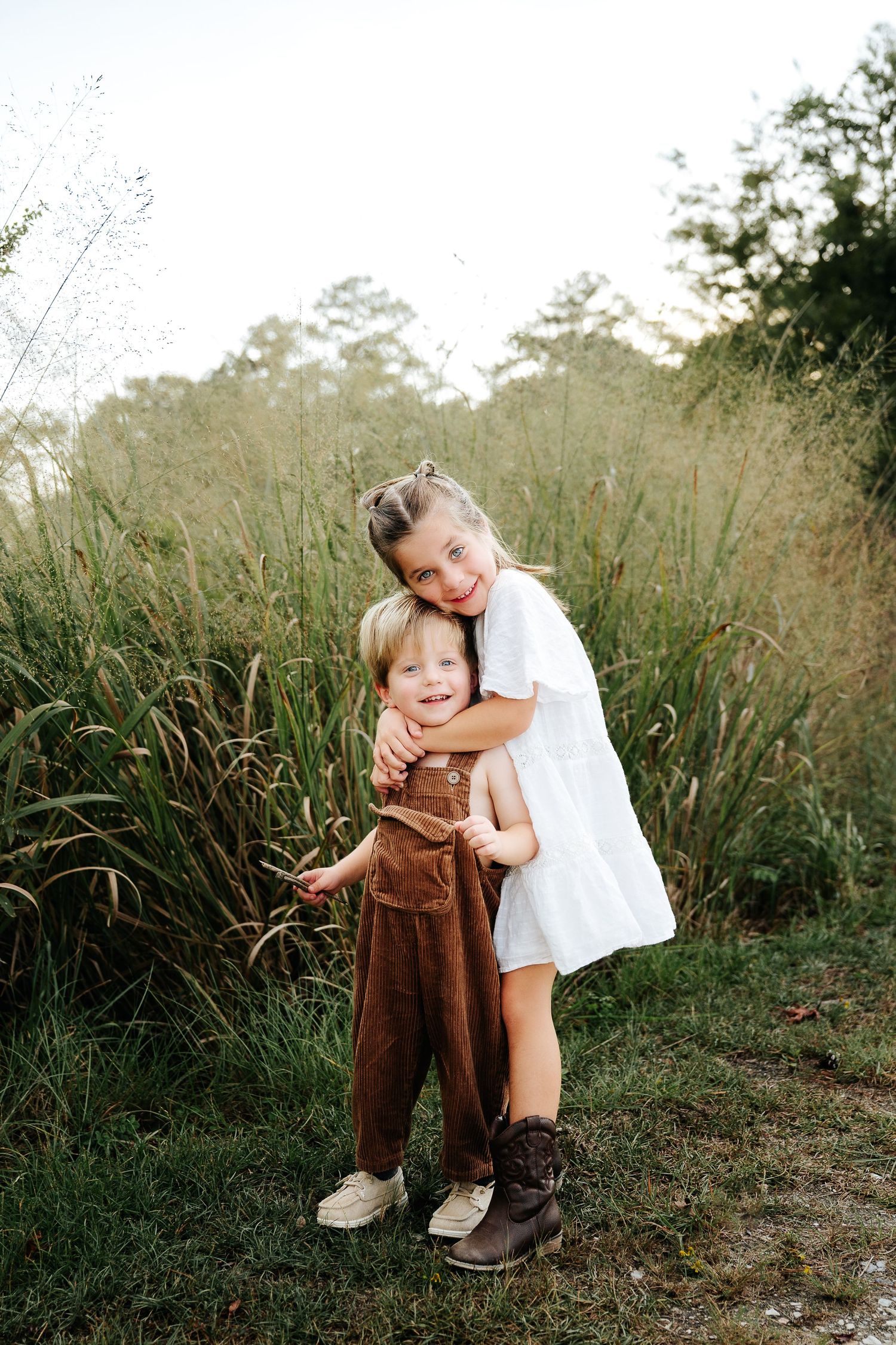 Two young siblings share a warm hug outdoors, girl in white dress embracing toddler in brown overalls near tall grass.