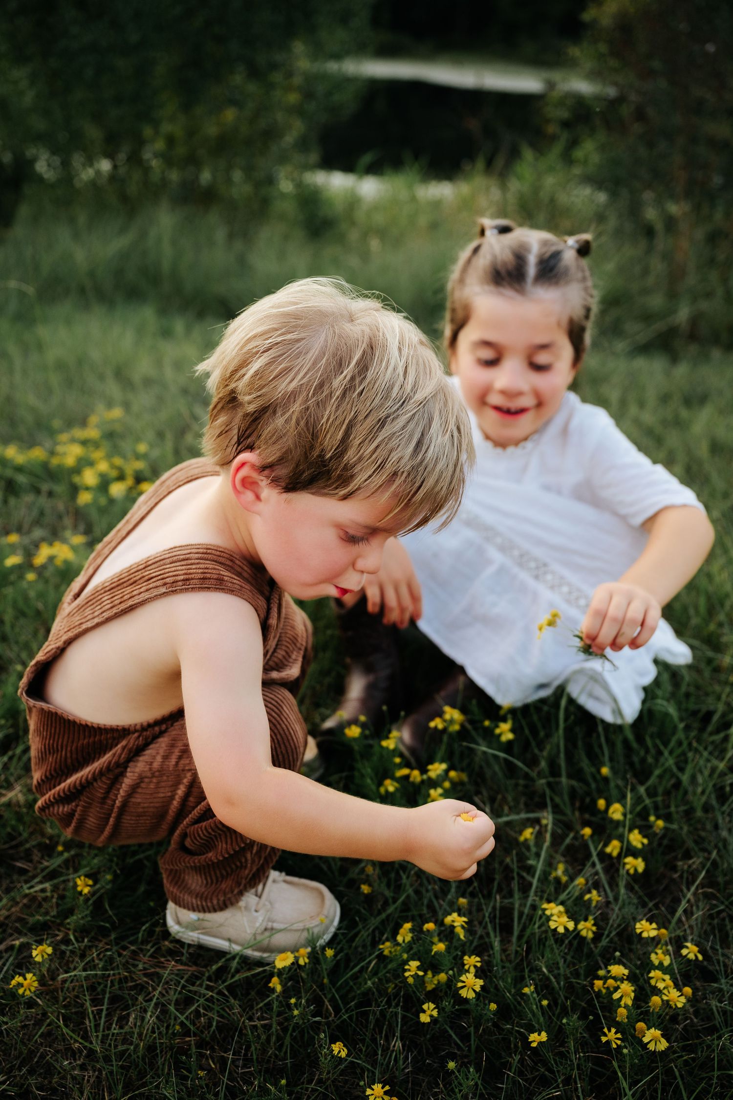 Two young children exploring yellow wildflowers in a green meadow, boy in brown romper crouching beside smiling girl in white dress.