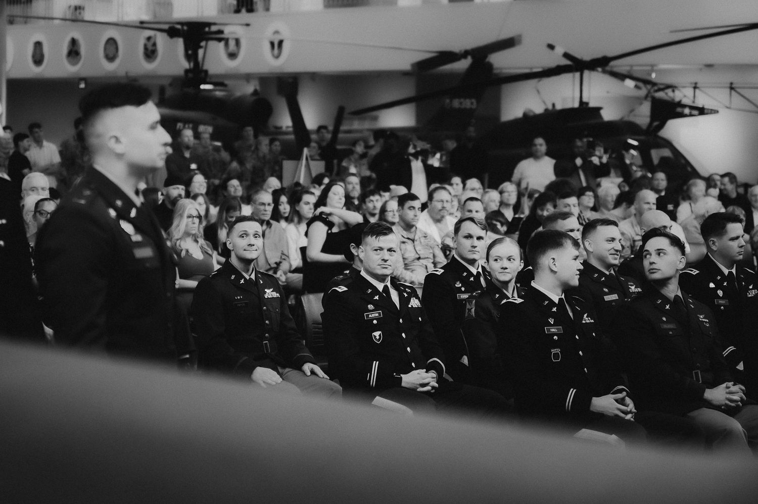 A black and white photograph shows a crowded audience sitting in rows of theater-style seating.
