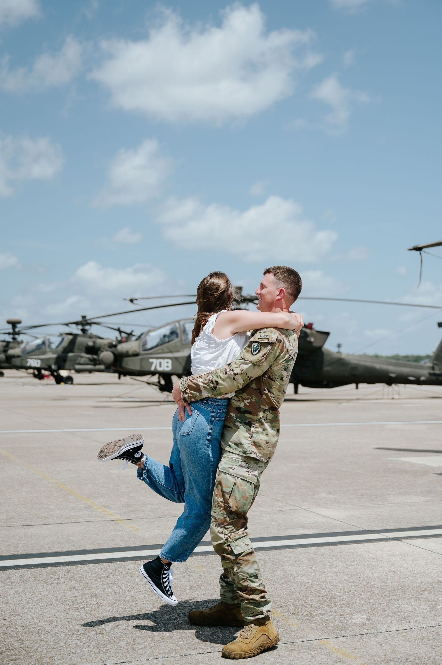 A soldier in camouflage uniform joyfully lifts a woman in jeans near military helicopters on a sunny airfield.