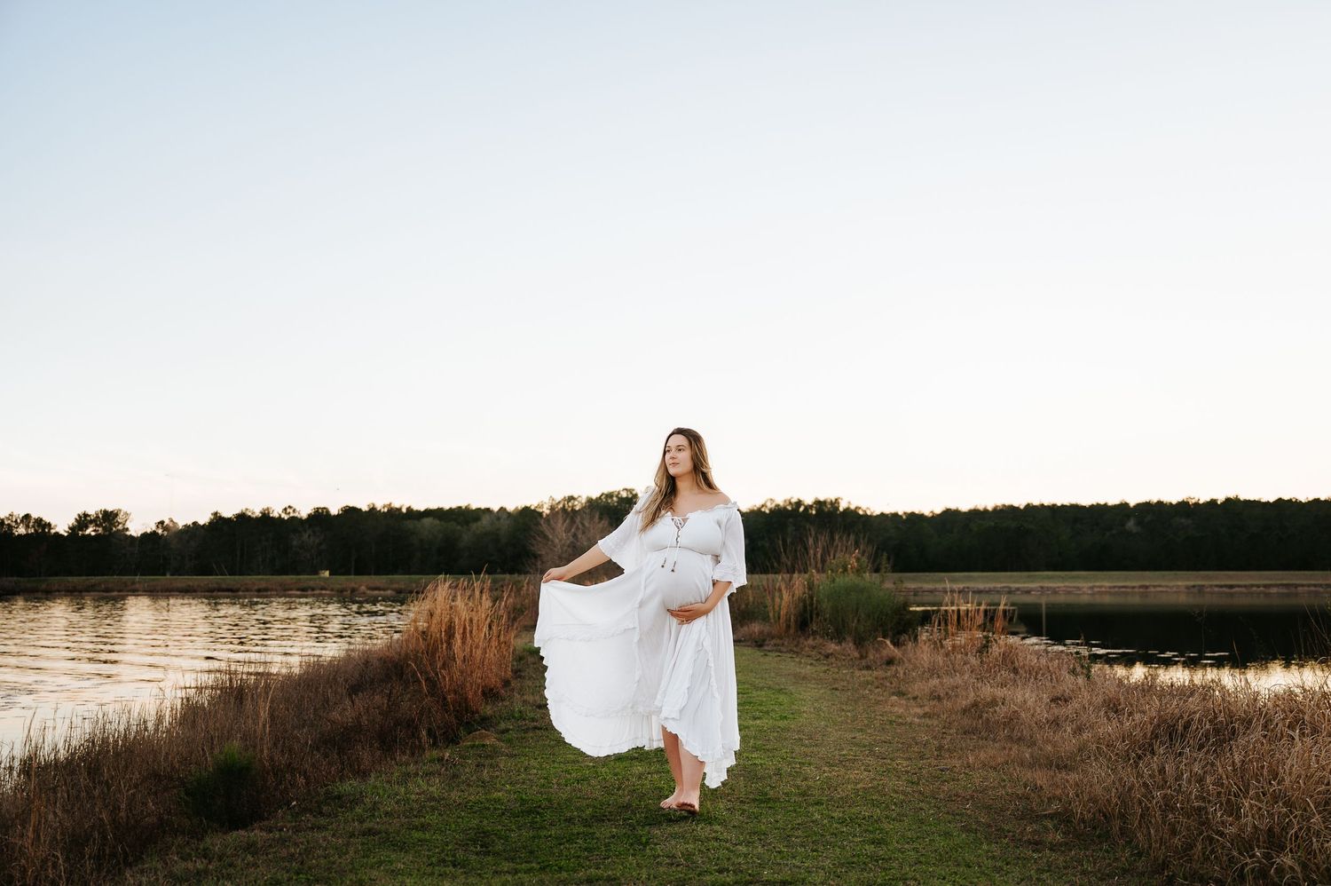 A figure in a flowing white dress stands gracefully in a scenic field at sunset near a serene lake.