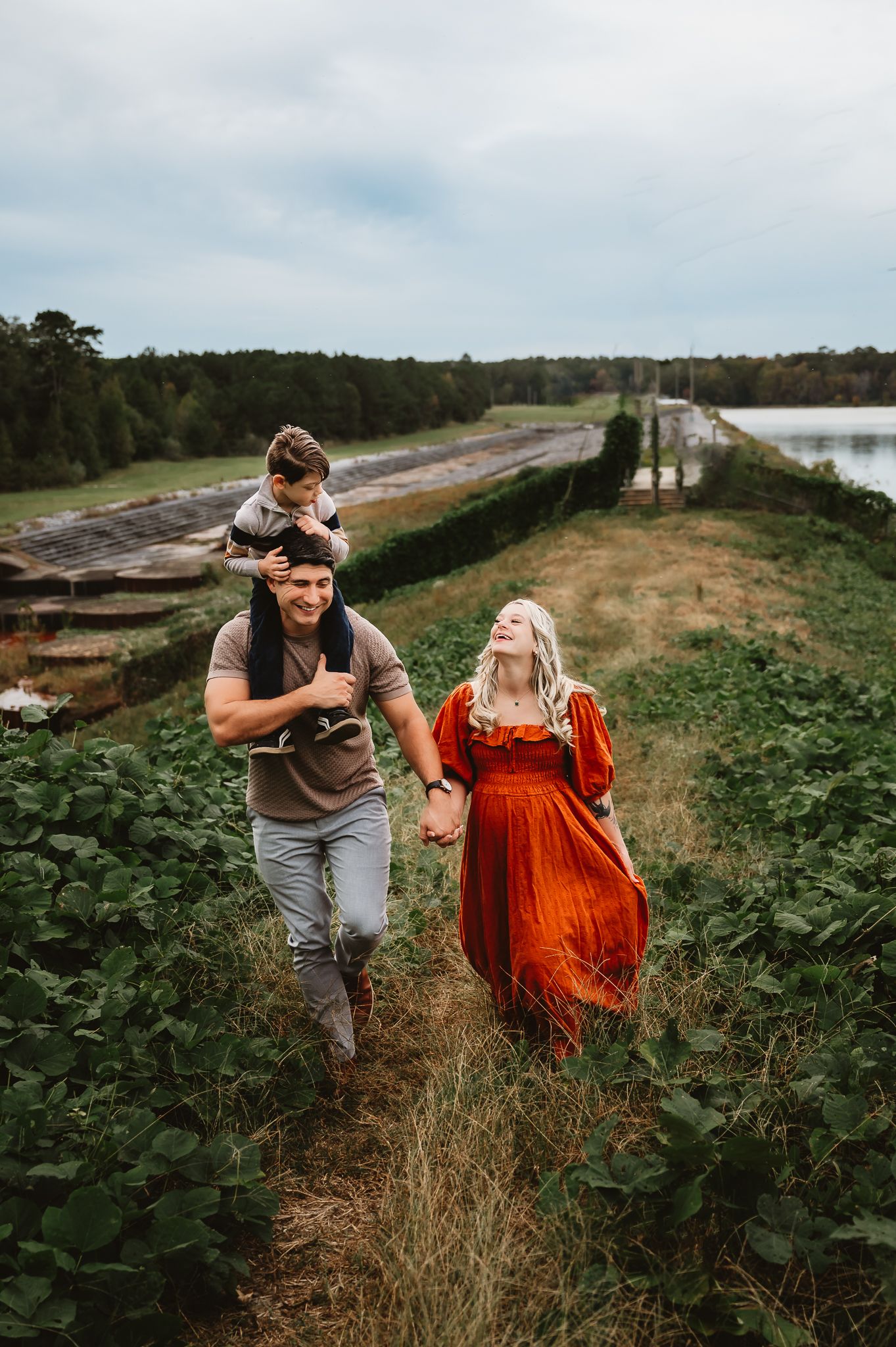 A couple walks together through tall grass and greenery along a waterfront path on a cloudy day.