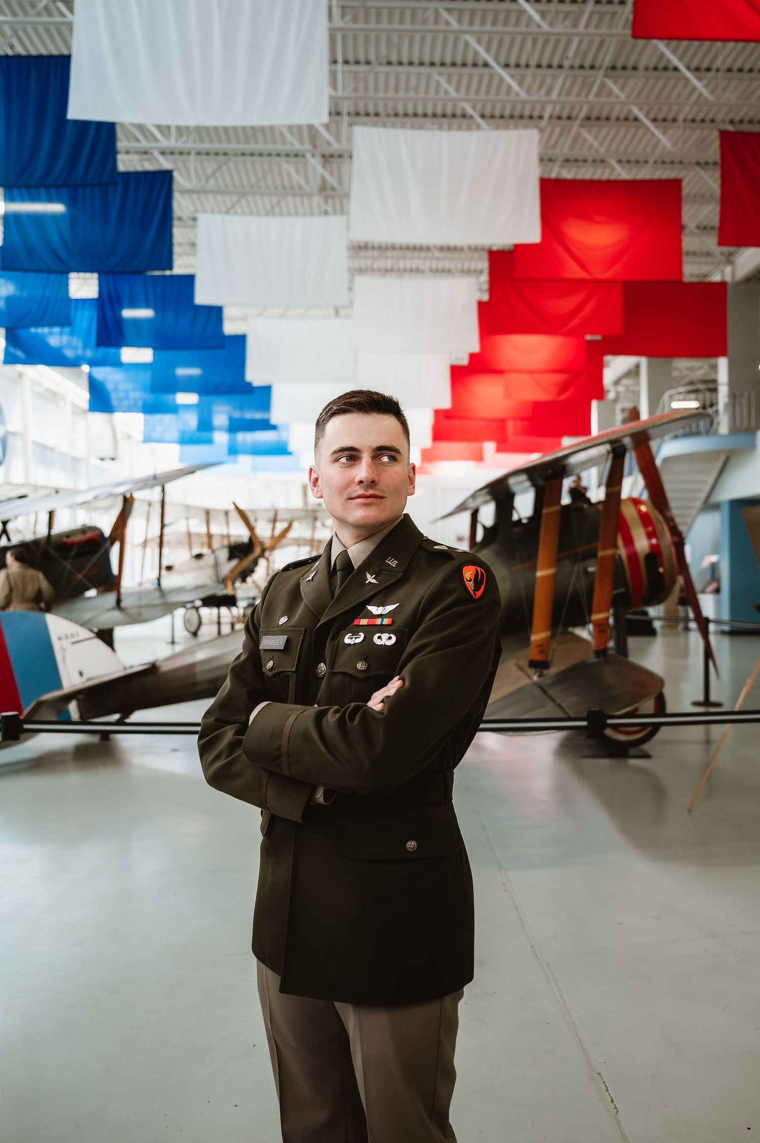 A young military officer in dress uniform stands with arms crossed in front of vintage aircraft in a museum with red, white, and blue flags.