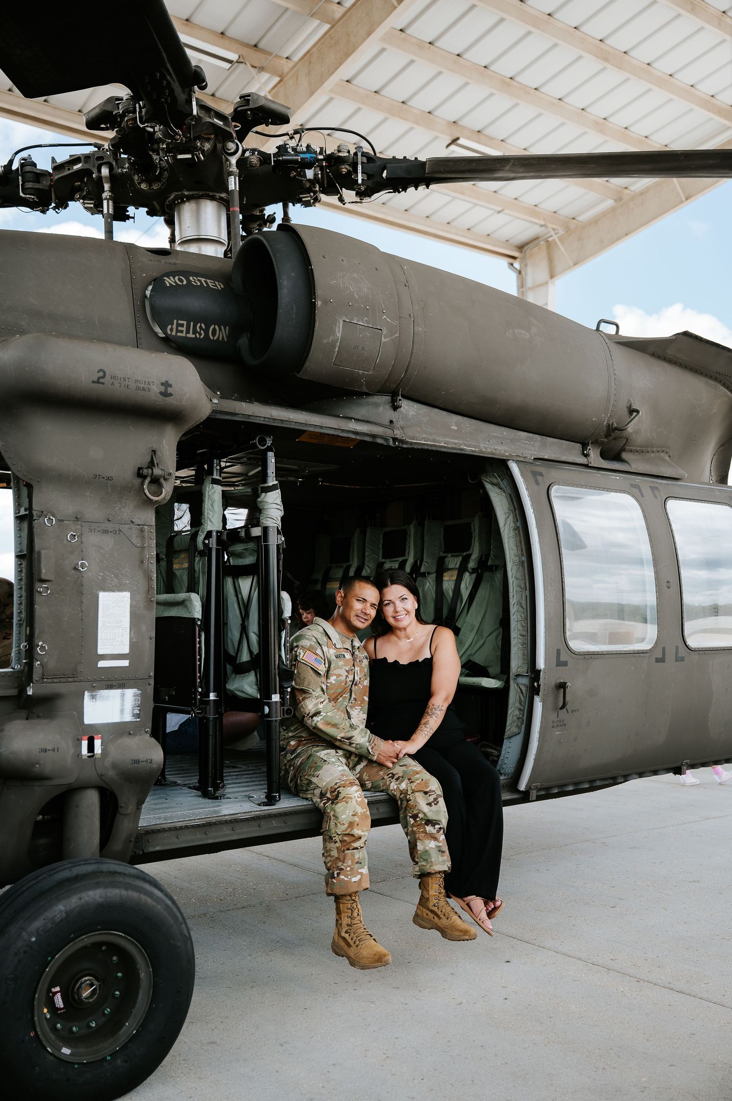 Military couple sitting together in open helicopter door at hangar, soldier in uniform embracing partner in black dress.