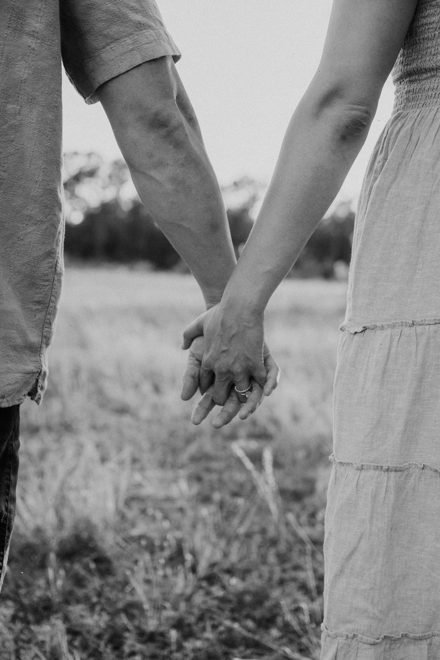 Black and white photo of a couple holding hands in a field, woman wearing a ring on her finger.