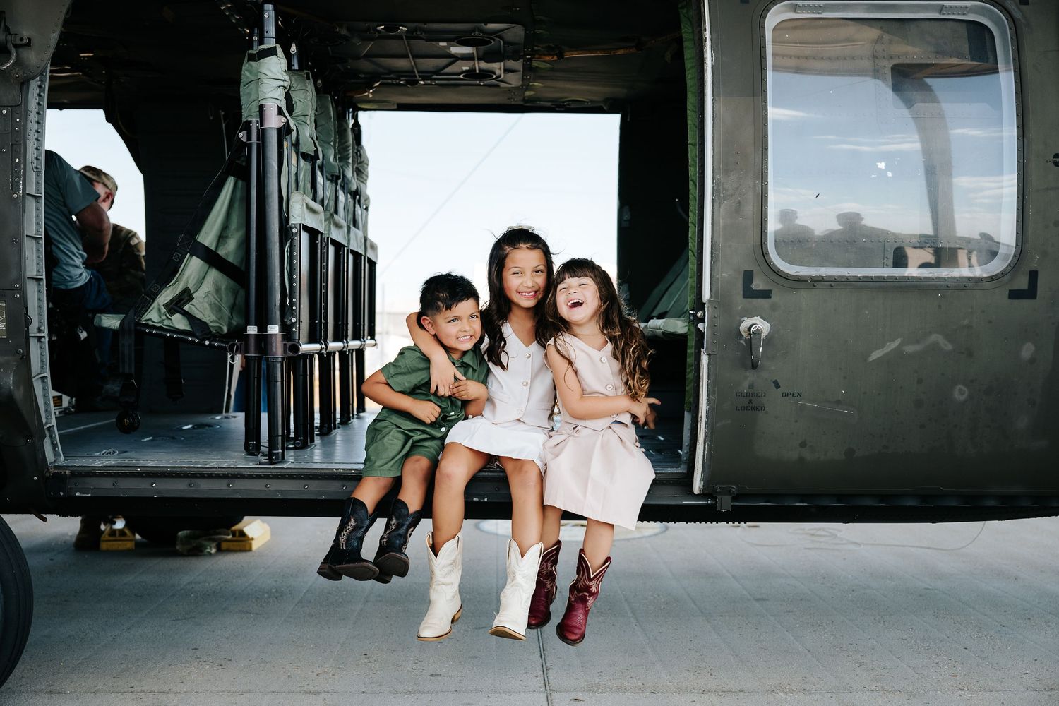 Three children sitting and smiling in the open door of a military helicopter on a tarmac.