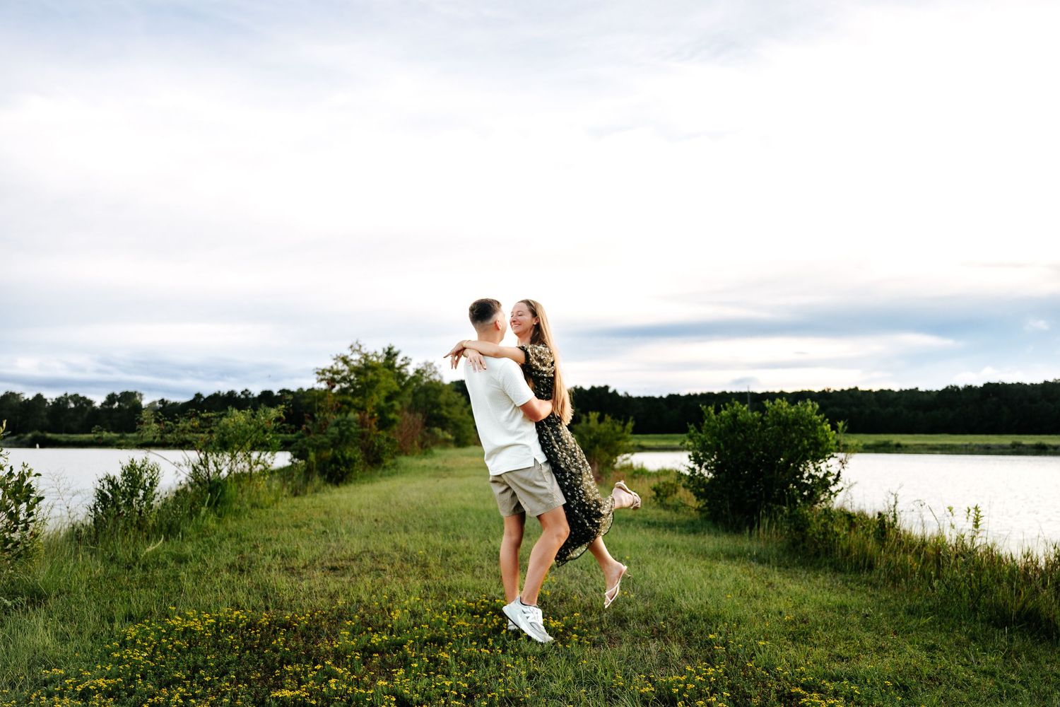 Couple embracing and kissing outdoors near a lake, surrounded by green grass and trees under a cloudy sky.