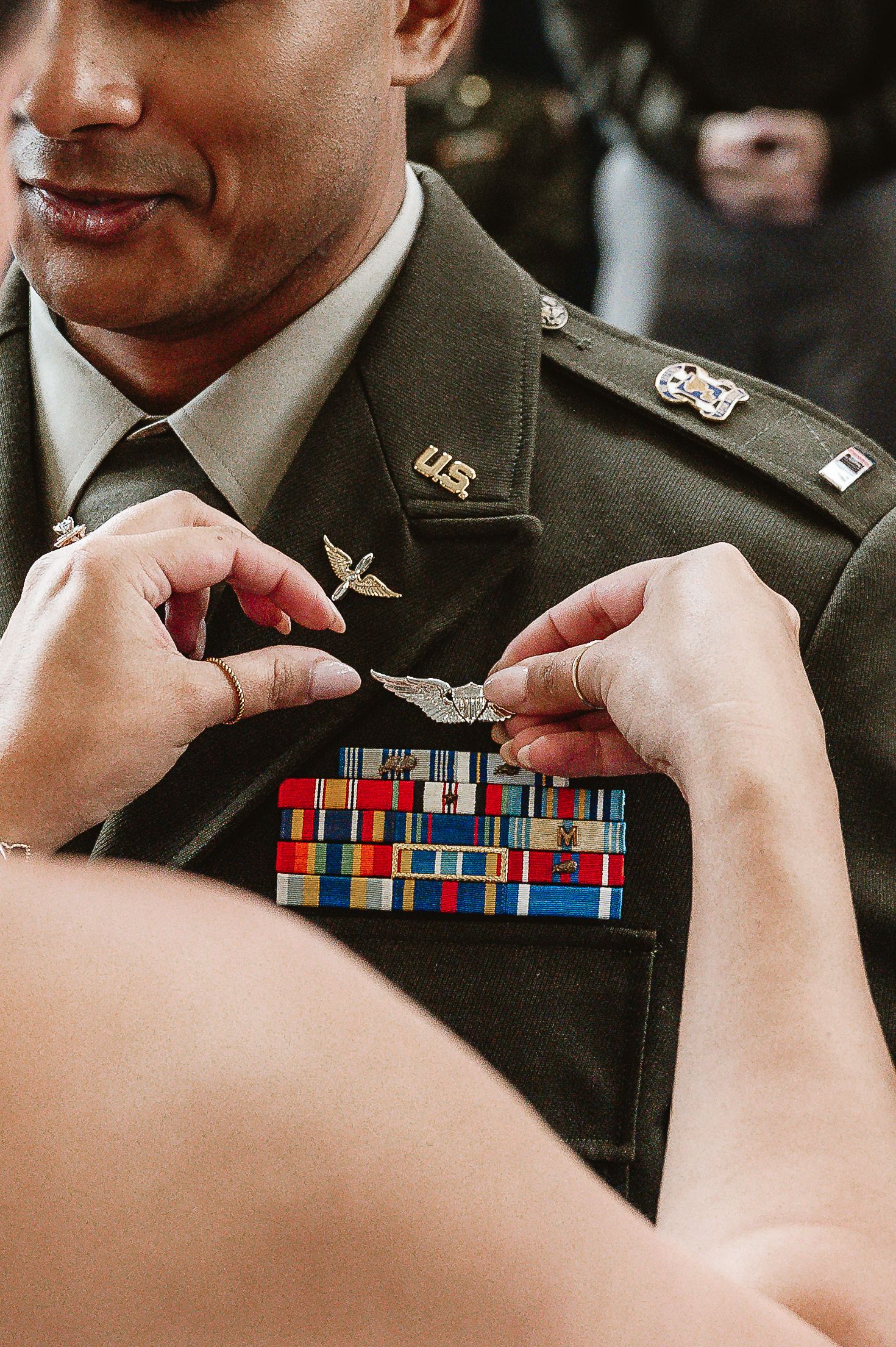 Hands pinning military aviator wings badge on U.S. Army dress uniform with colorful service ribbons.