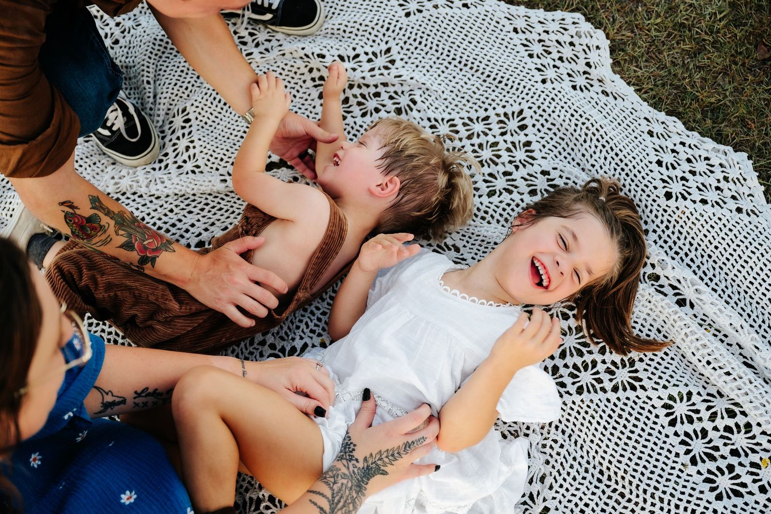 Two laughing children play on a white crochet blanket outdoors while a tattooed adult watches over them.