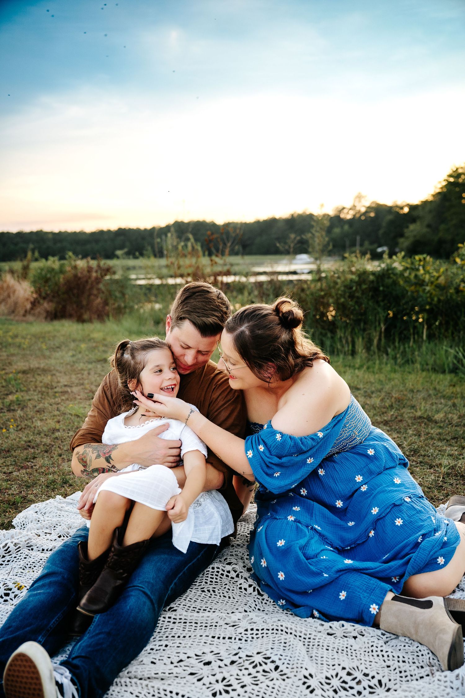 A happy family sitting on a blanket outdoors, parents kissing their laughing young daughter near a scenic lake.