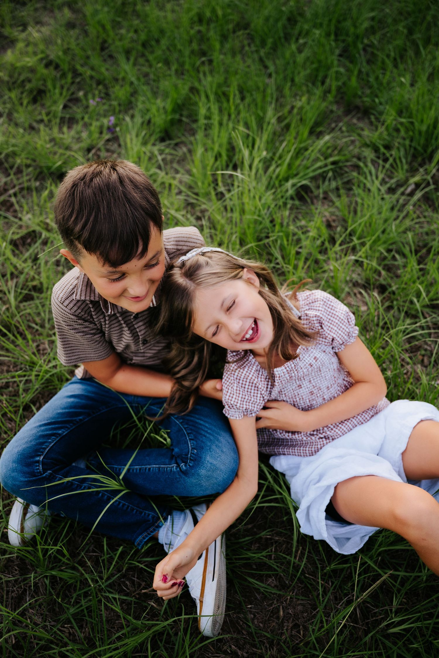 Two happy children sitting in green grass, laughing and hugging outdoors on a sunny day.