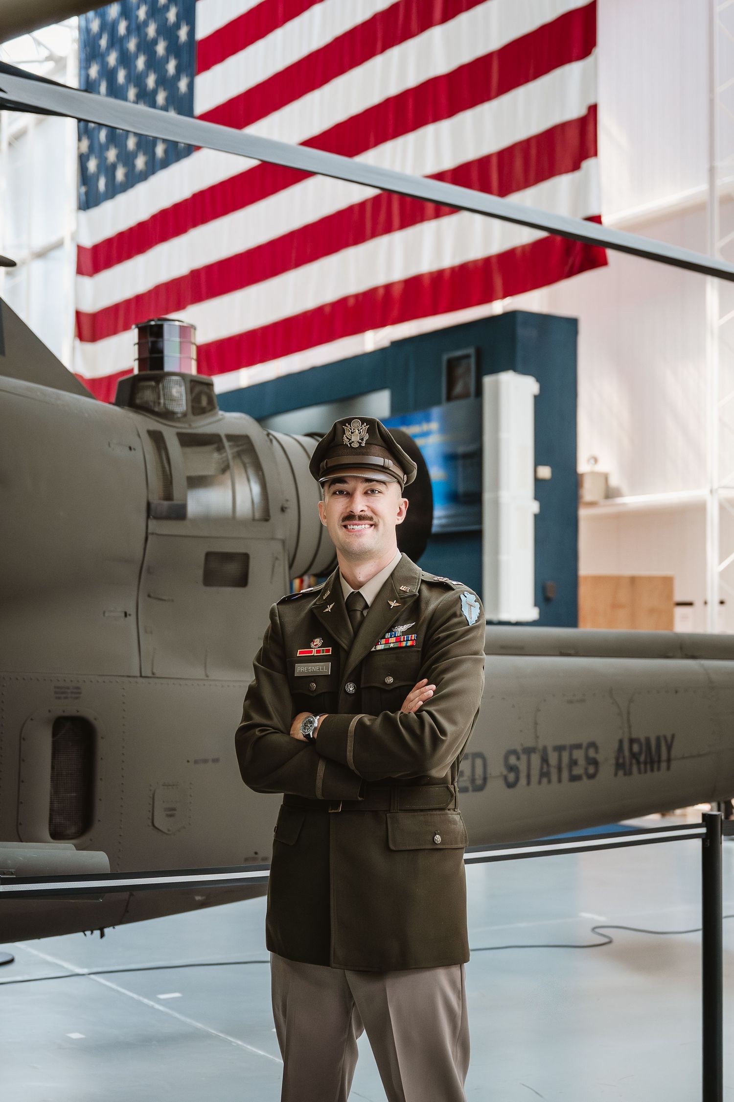 US Army officer in dress uniform stands arms crossed before a military helicopter and American flag display.