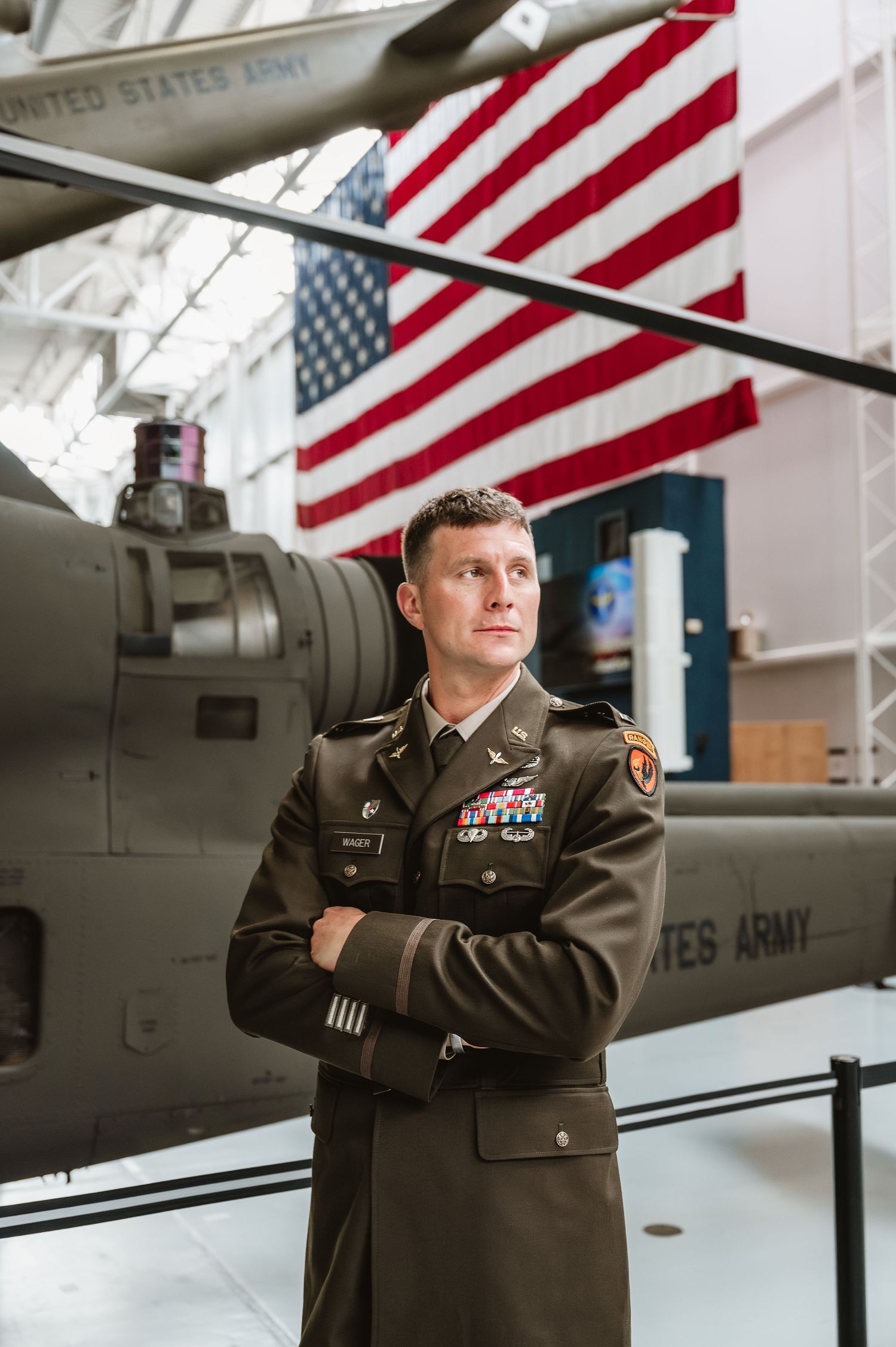 US Army officer in dress uniform stands arms crossed before a military helicopter and American flag display.
