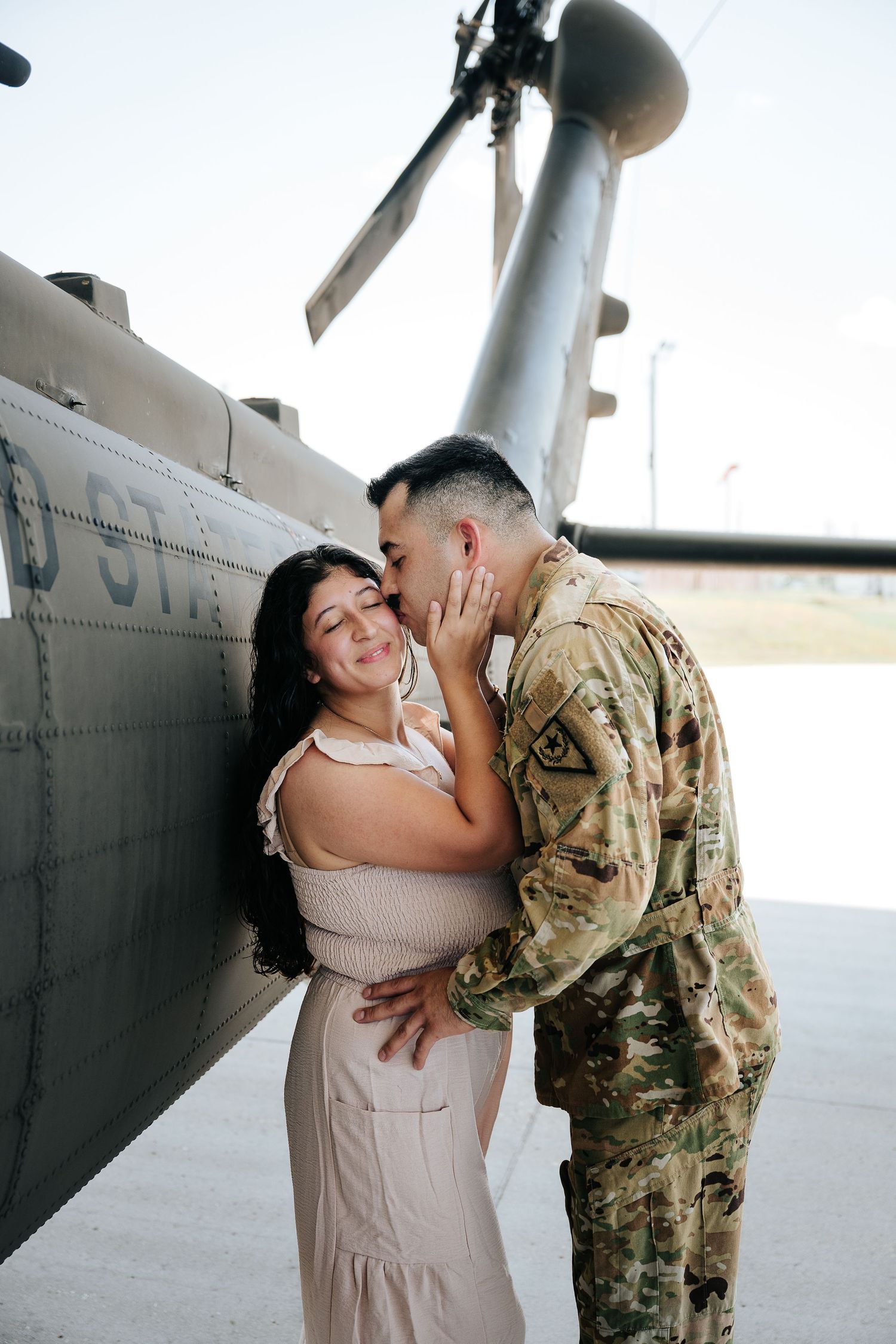A soldier in camouflage uniform kisses a woman in a pink dress beside a military helicopter.