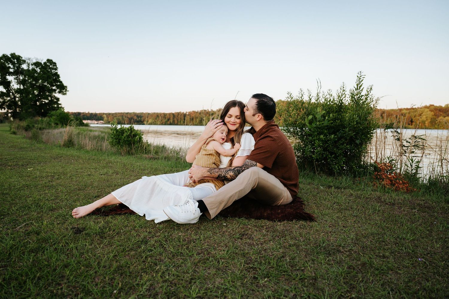 Couple sitting together on grass by a serene lake at sunset, sharing a tender moment outdoors.