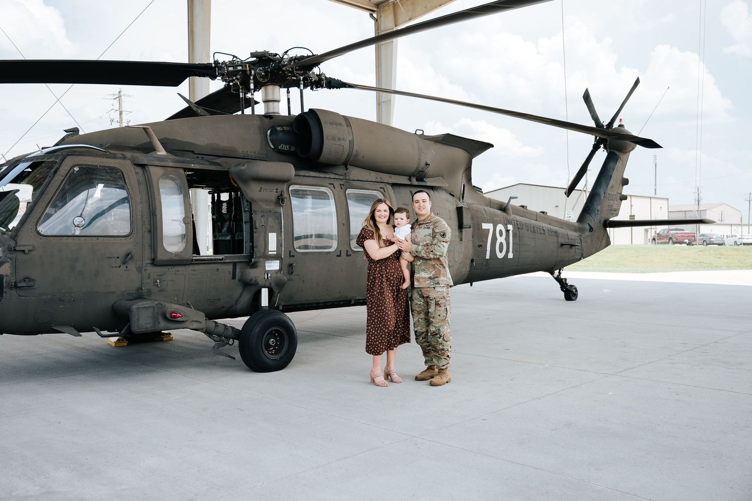 Couple in military uniform poses with baby in front of UH-60 Black Hawk helicopter numbered 781 at airfield.