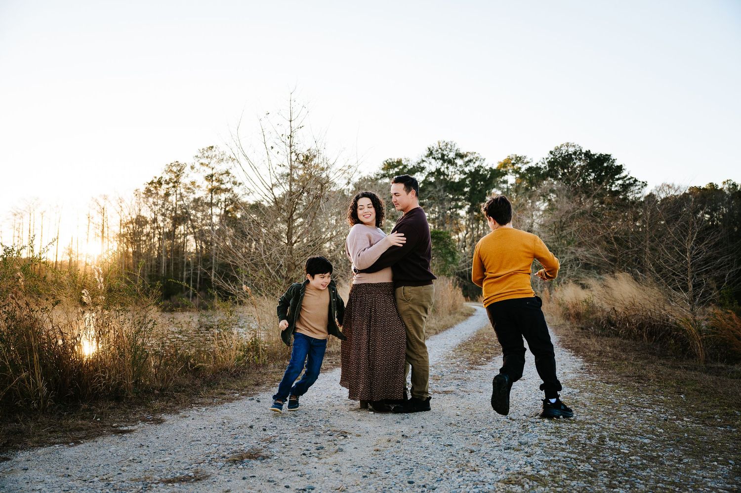 Family enjoying a sunset walk together on a dirt path surrounded by tall grass and trees near a lake.