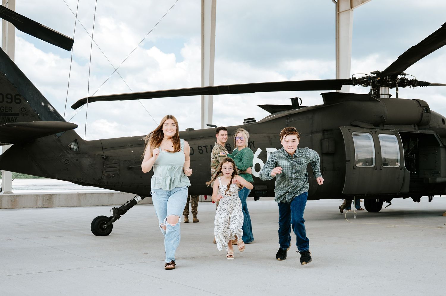 Military family runs toward camera in front of a Black Hawk helicopter on a rooftop during a photo session.