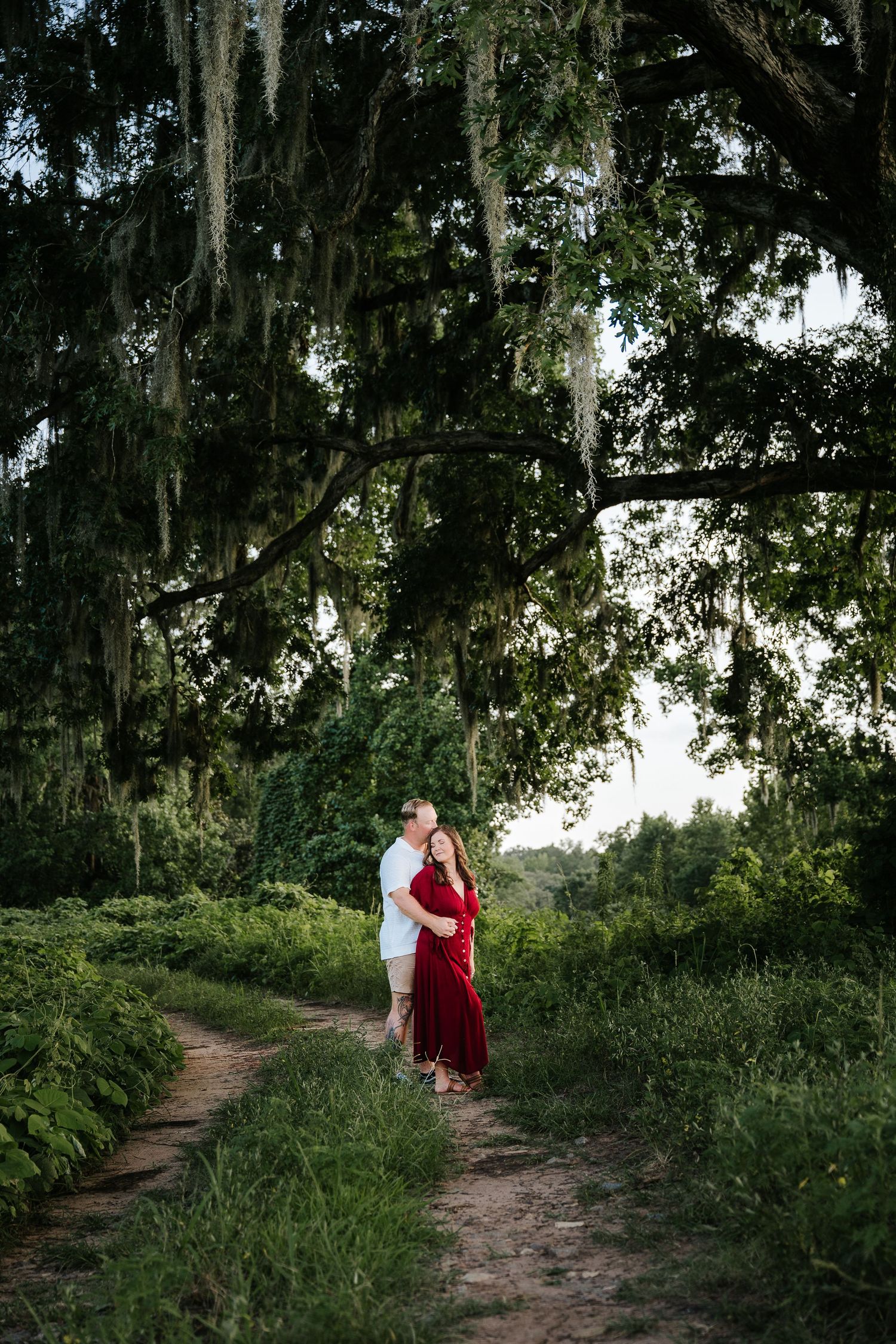 Couple embraces on a dirt path under a large moss-draped oak tree, woman in red dress, lush green surroundings.