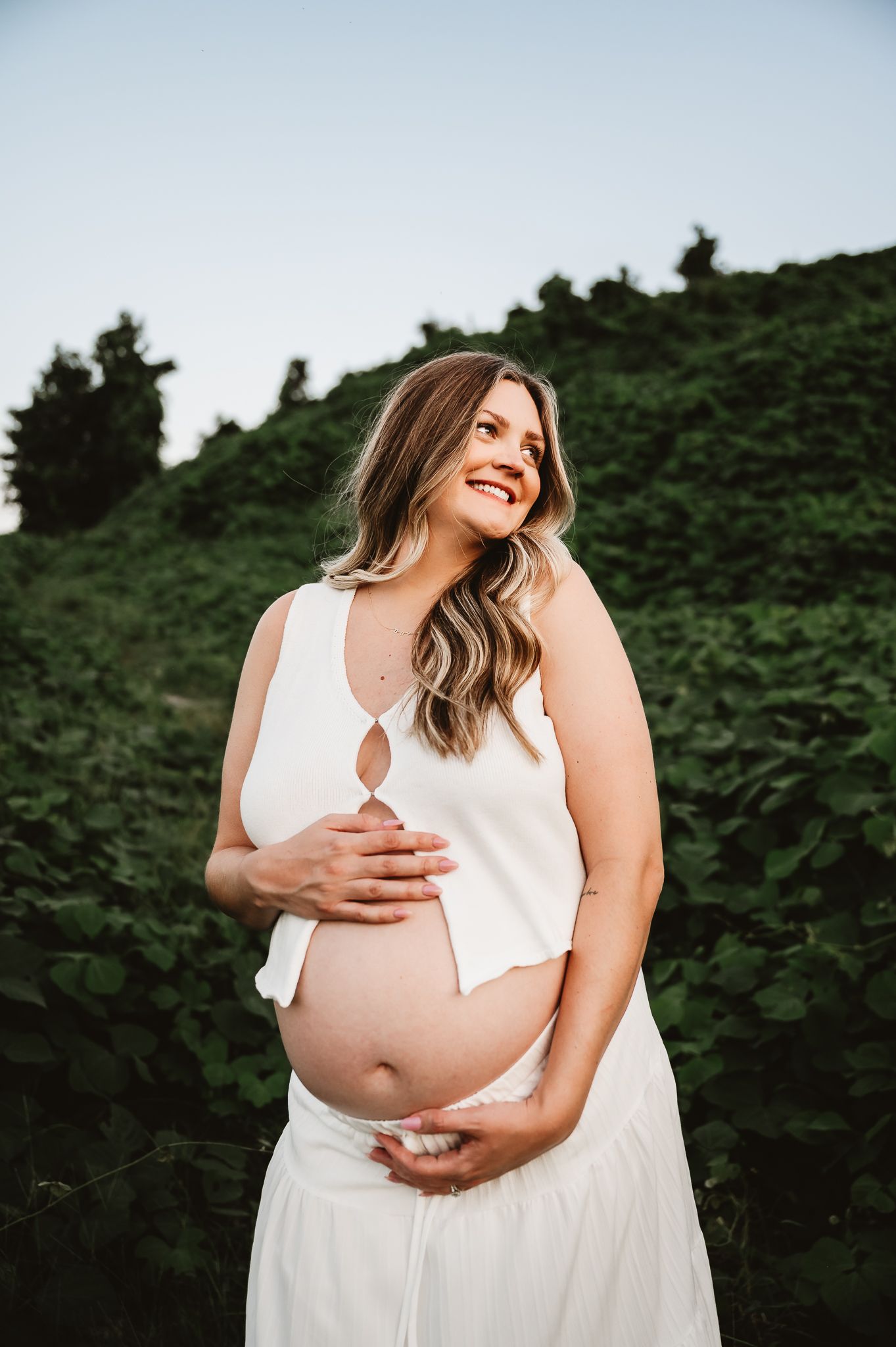 A maternity photoshoot featuring an expecting mother in a white dress outdoors against a backdrop of green foliage.