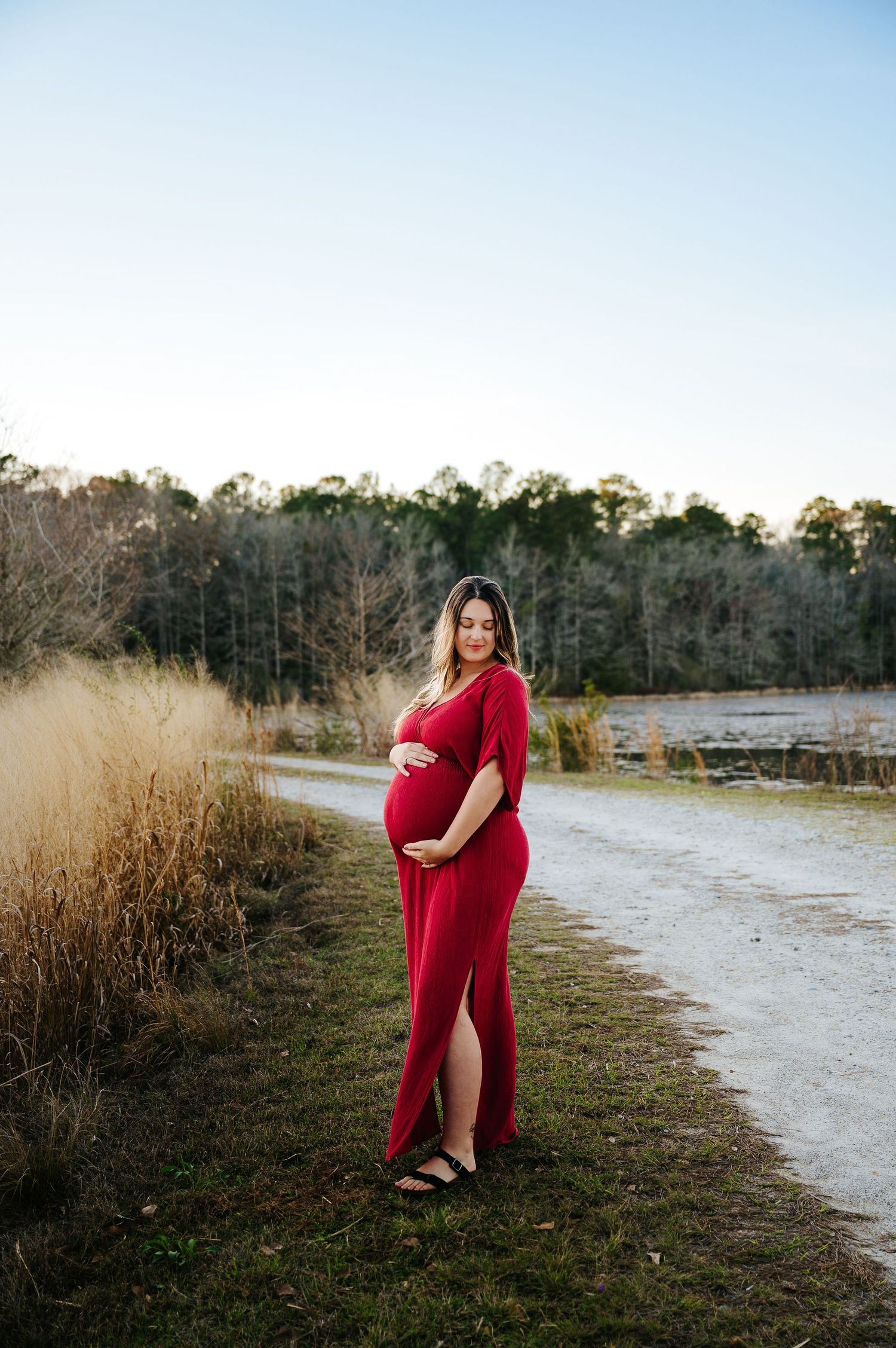 Pregnant woman in red maternity gown poses outdoors on a path surrounded by golden grass and trees.