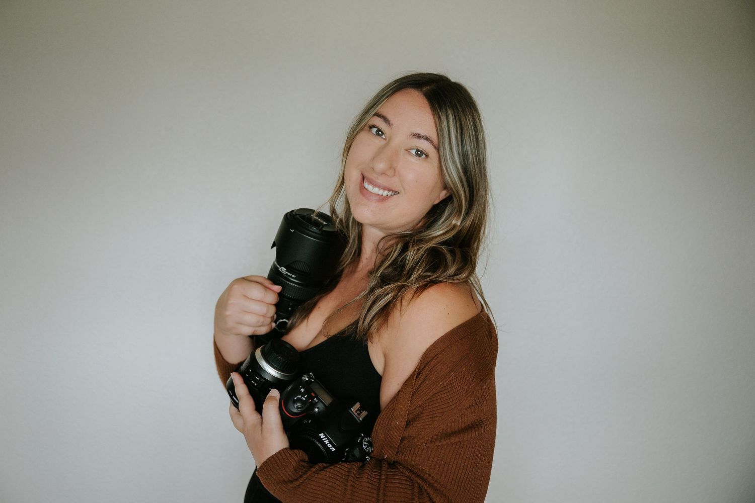 Smiling woman with highlighted hair holding a professional camera, wearing a brown cardigan against a neutral background.